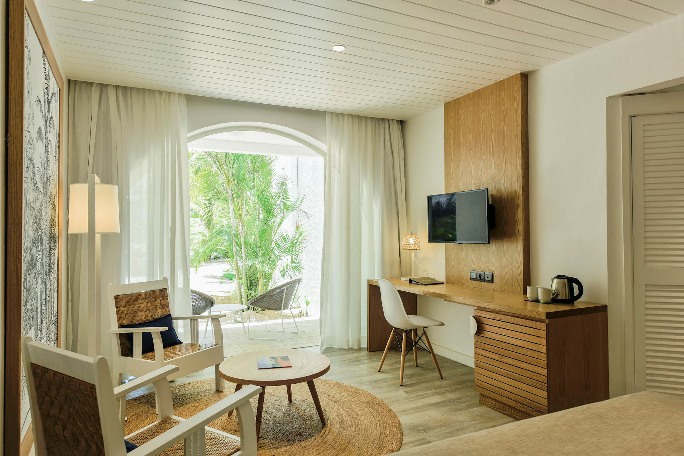 Ett modernt hotellrum med sittgrupp, skrivbord och en TV på väggen. En terrass i bakgrunden med utsikt över gröna växter. (AI-genererad text)