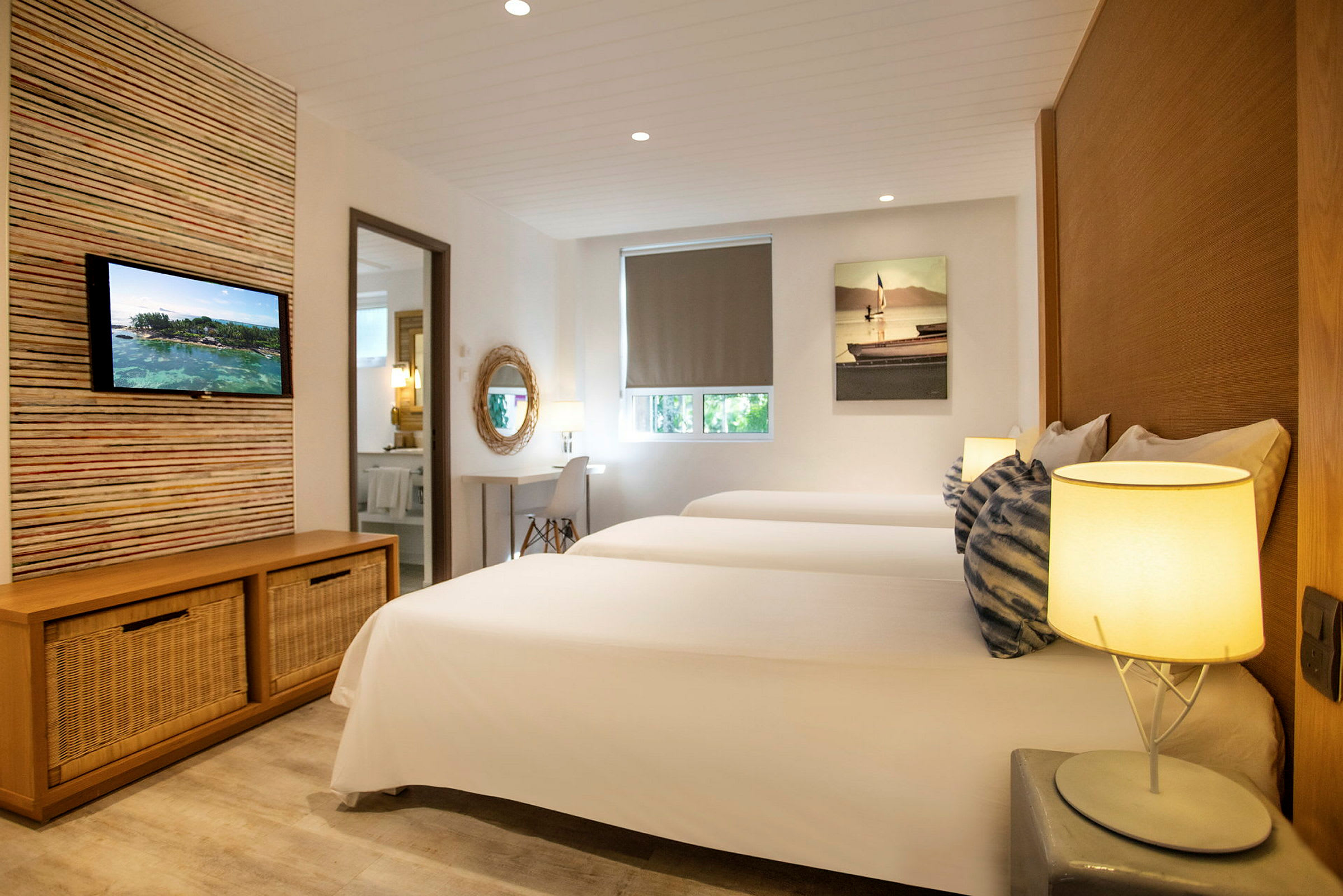 Ett hotellrum med tre vita sängar, en TV monterad på väggen, en skrivbordshörna och subtil belysning. (AI-genererad text)