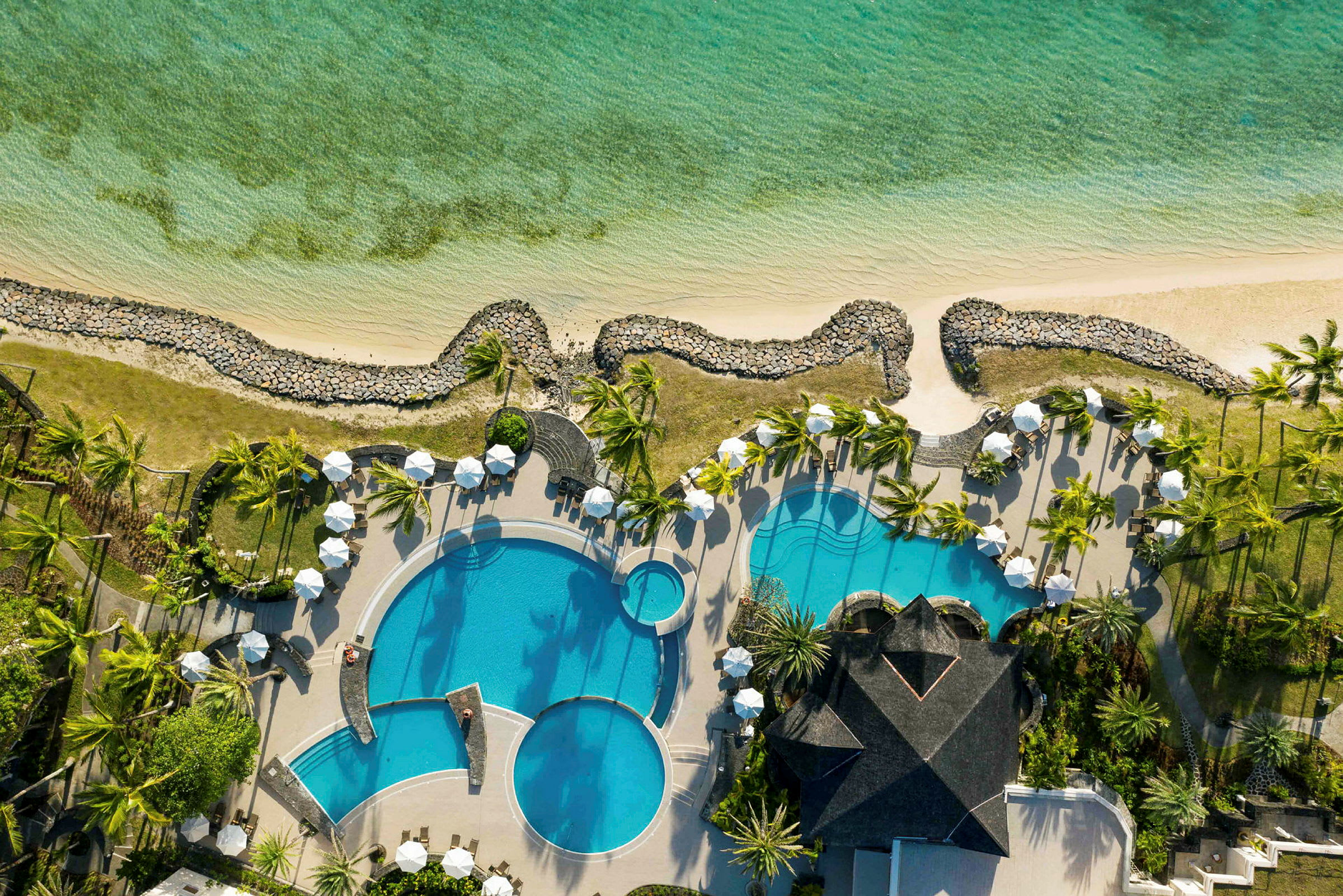 Flygfoto av ett hotellområde med pooler, solstolar och parasoller, omringat av grönska nära en kustlinje. (AI-genererad text)