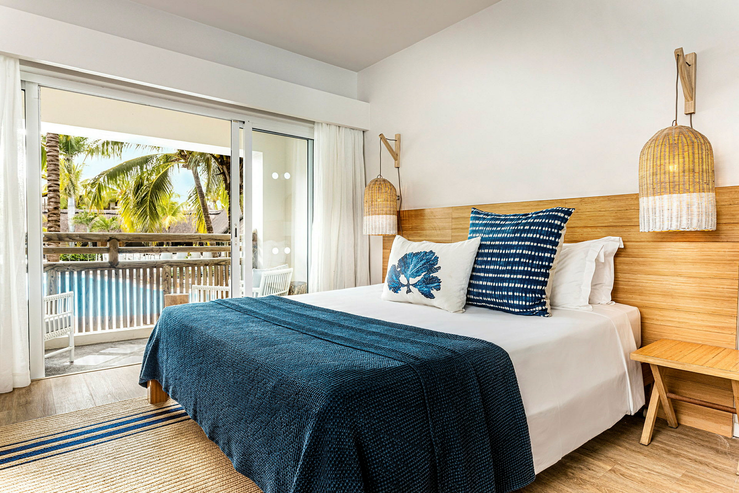 Ett hotellrum med en säng med blått överkast och dekorativa kuddar. Rummet har en balkong med utsikt över en pool och palmer. (AI-genererad text)