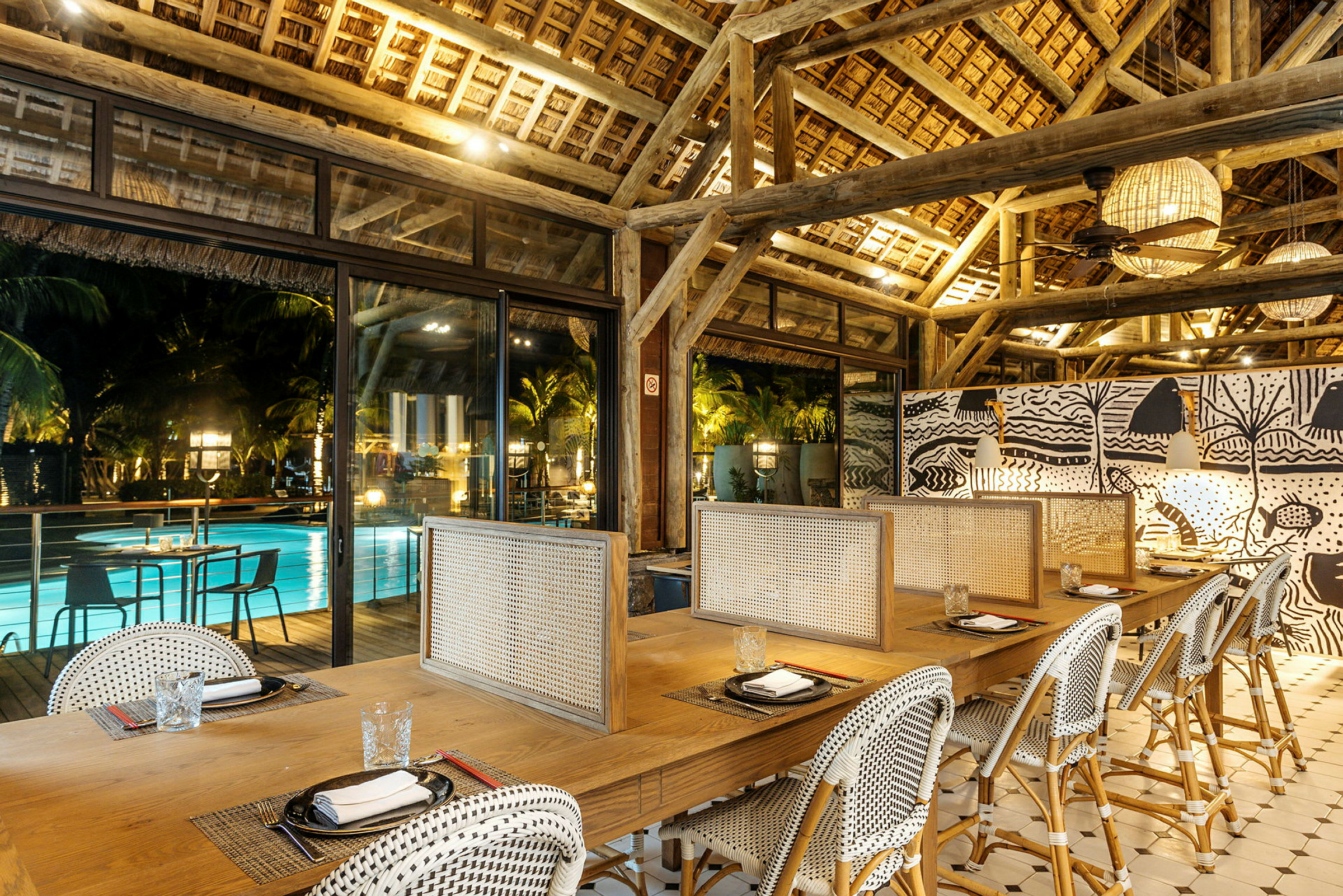 En restaurang med bord och stolar framför stora fönster som vetter mot en upplyst pool och palmer. (AI-genererad text)