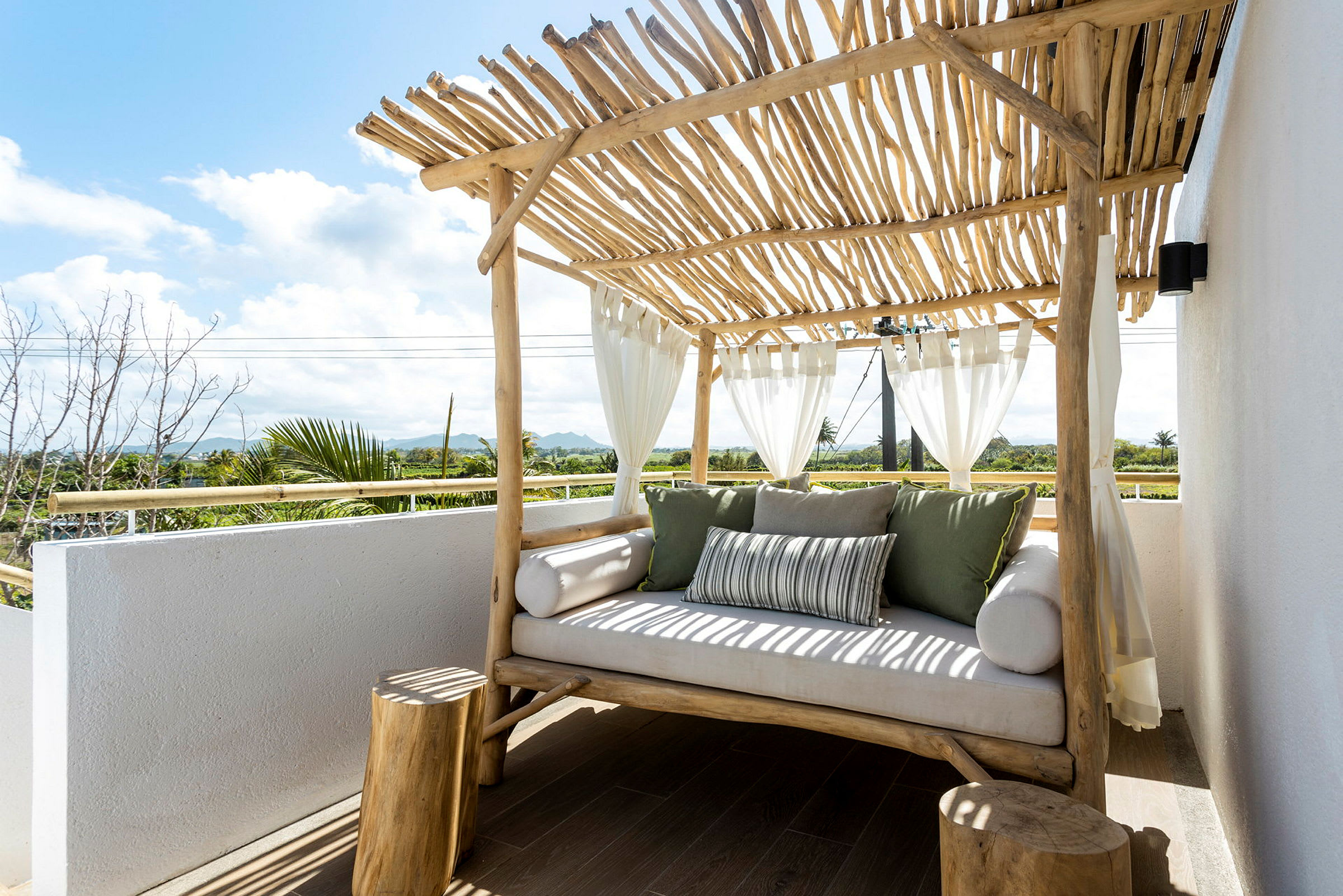 Vit soffa med gröna och randiga kuddar under ett tak av trästavar, med utsikt över grönska och en blå himmel. (AI-genererad text)