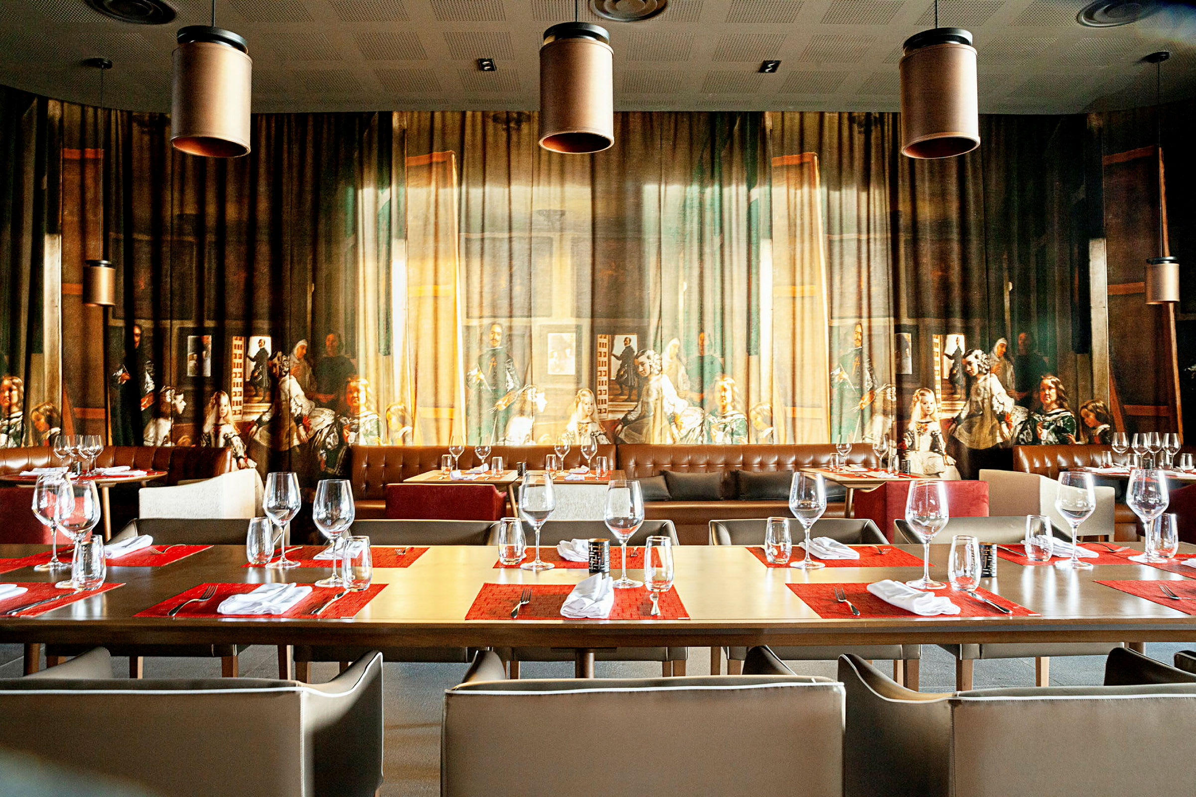 En restaurang med bord dukade med glas och servetter framför en vägg med ett stort, detaljrikt konstverk. (AI-genererad text)
