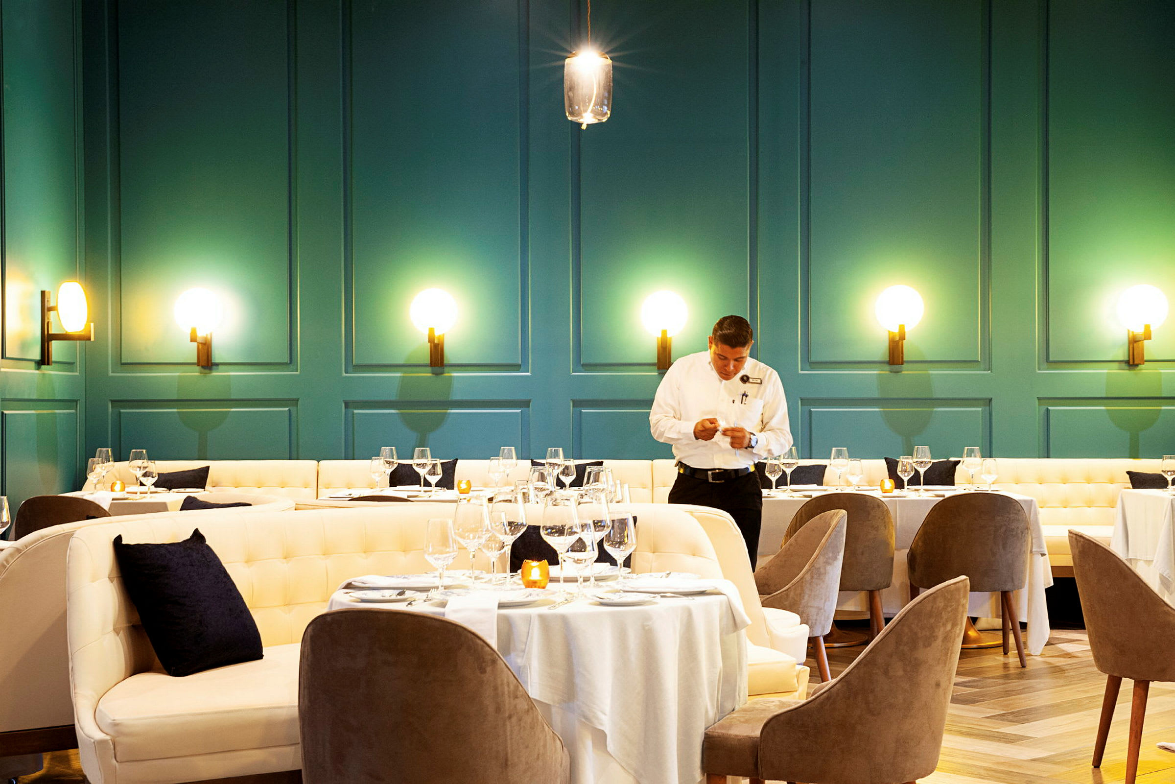 En elegant serveringslokal med dukade matbord omgivna av stoppade stolar och soffor. Väggarna är målade i en djup grön färg och pryds av moderna lampor. (AI-genererad text)