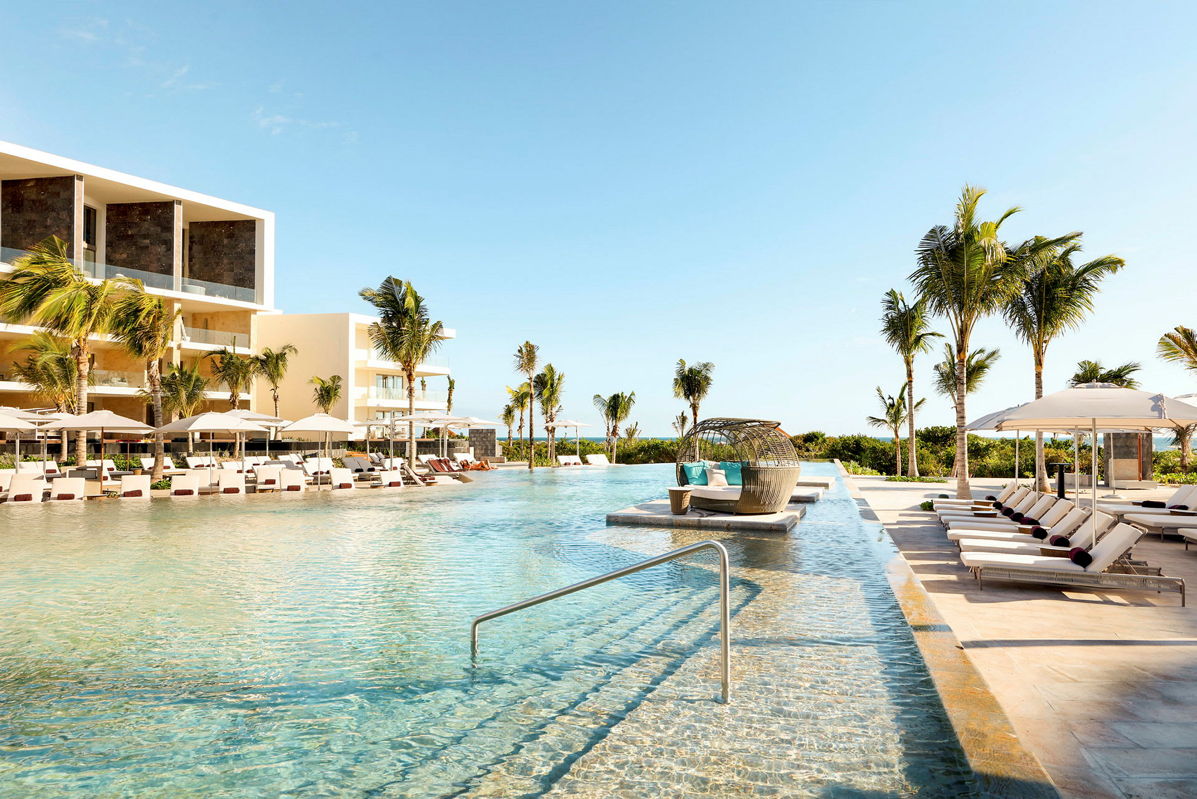 En stor pool med solstolar och parasoller vid kanten, omgiven av palmer och moderna hotellbyggnader. (AI-genererad text)