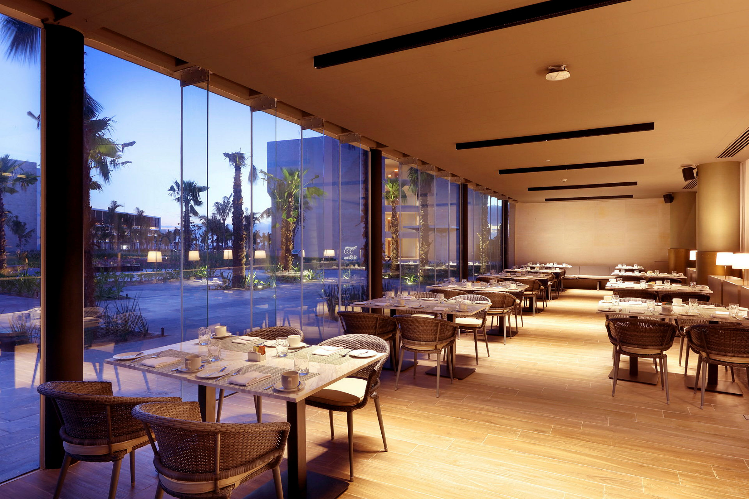 En modern restaurang med dukade bord och glasväggar som vetter mot en utomhusmiljö med palmer och byggnader. (AI-genererad text)