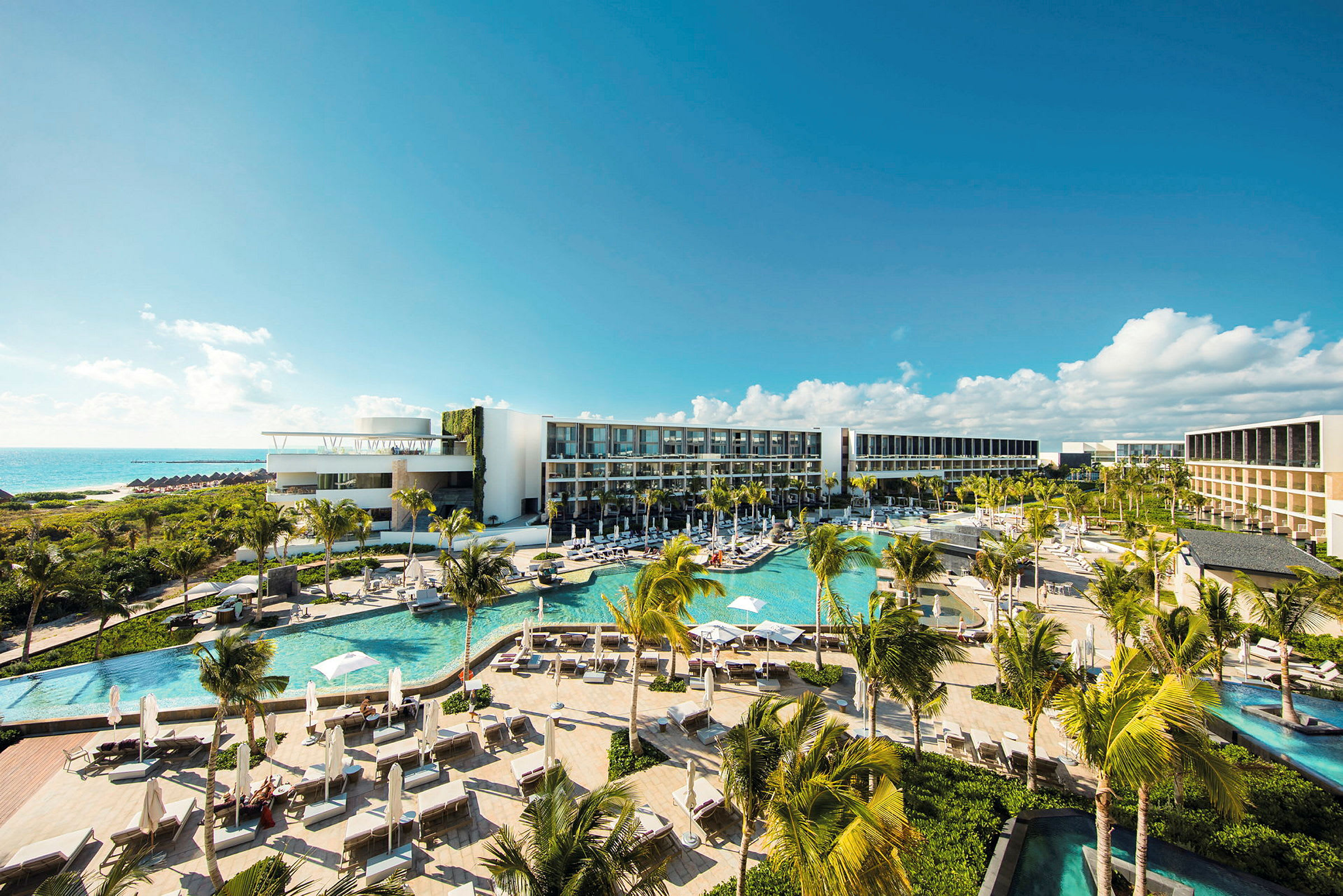 Ett modernt hotellkomplex med flera pooler, frodig vegetation och palmer, beläget vid kusten med utsikt över havet under en klarblå himmel. (AI-genererad text)