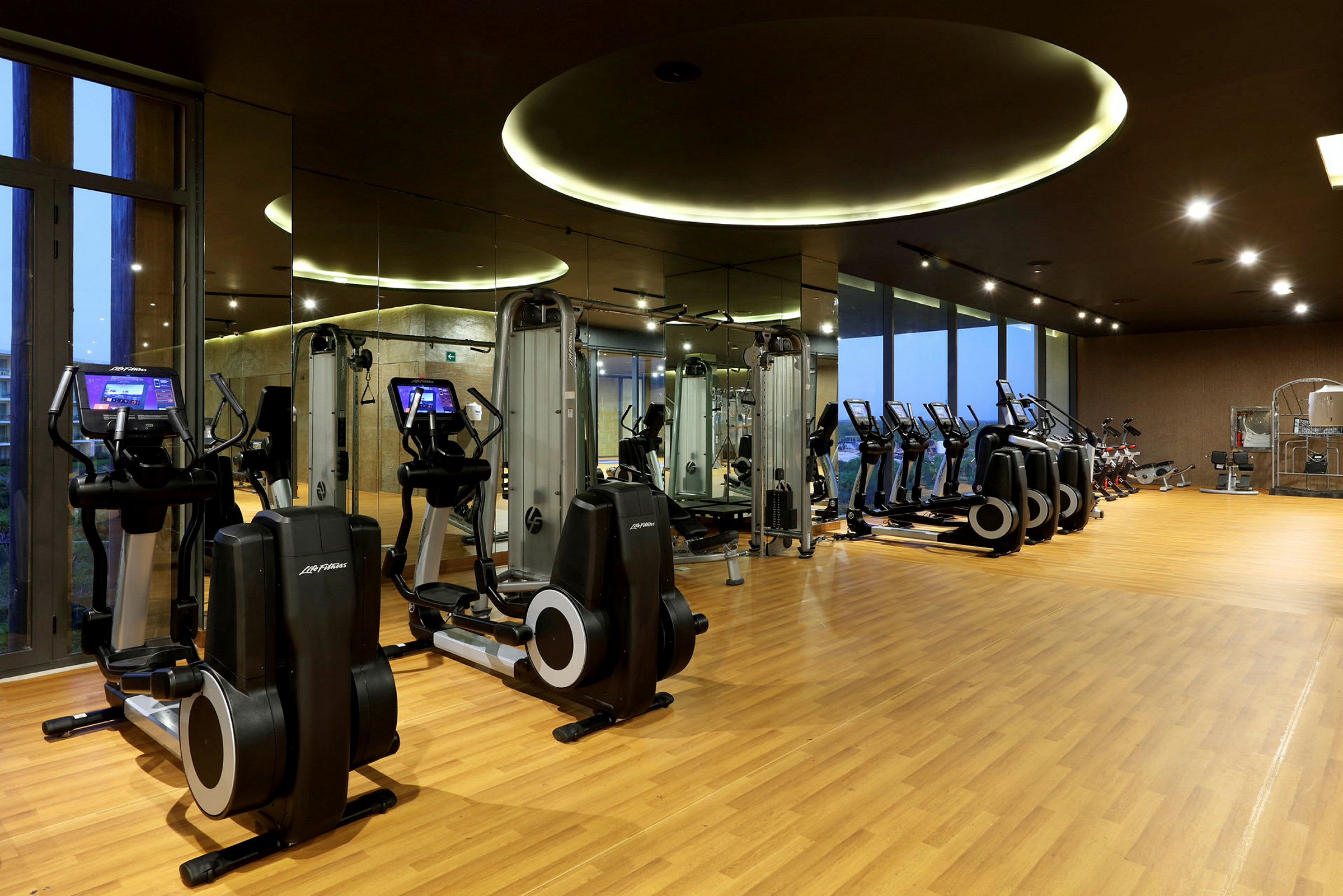 Ett gym med flera träningsmaskiner, inklusive löpband, crosstrainers och styrketräningsutrustning. Lokalen har trägolv och speglar längs ena väggen. (AI-genererad text)