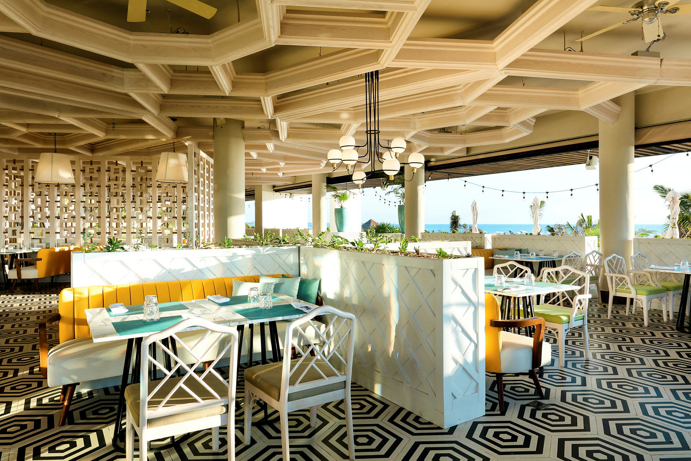 En modern restaurang med geometriskt mönstrat golv, ljusa möbler och stora panoramafönster med utsikt mot havet. (AI-genererad text)