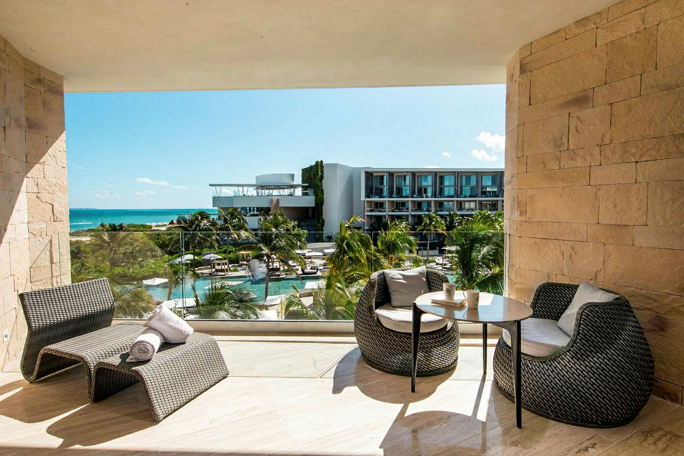 Utsikt från en balkong med modern utemöblering mot en poolanläggning med solstolar, följt av en byggnad och havet i bakgrunden. (AI-genererad text)