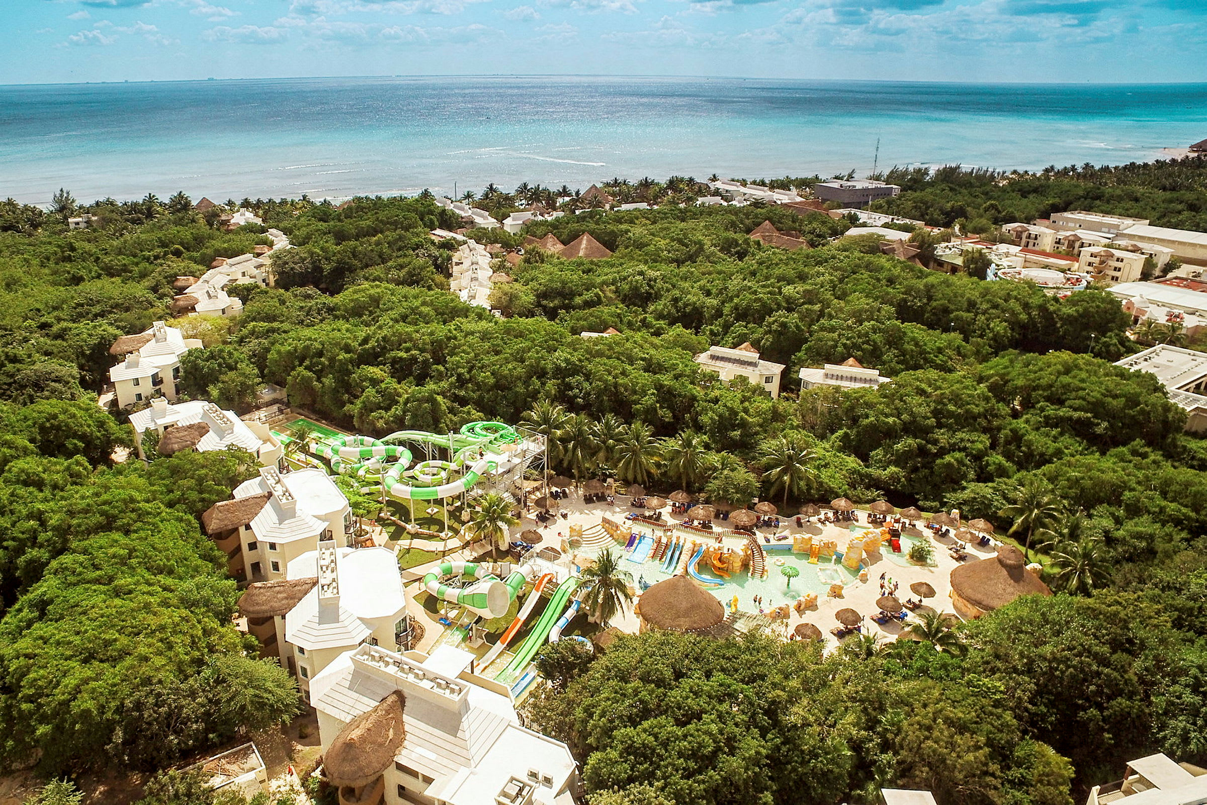 Flygfoto över en resort med pool- och vattenrutschområdet omgivet av grönska, med havet i bakgrunden. (AI-genererad text)