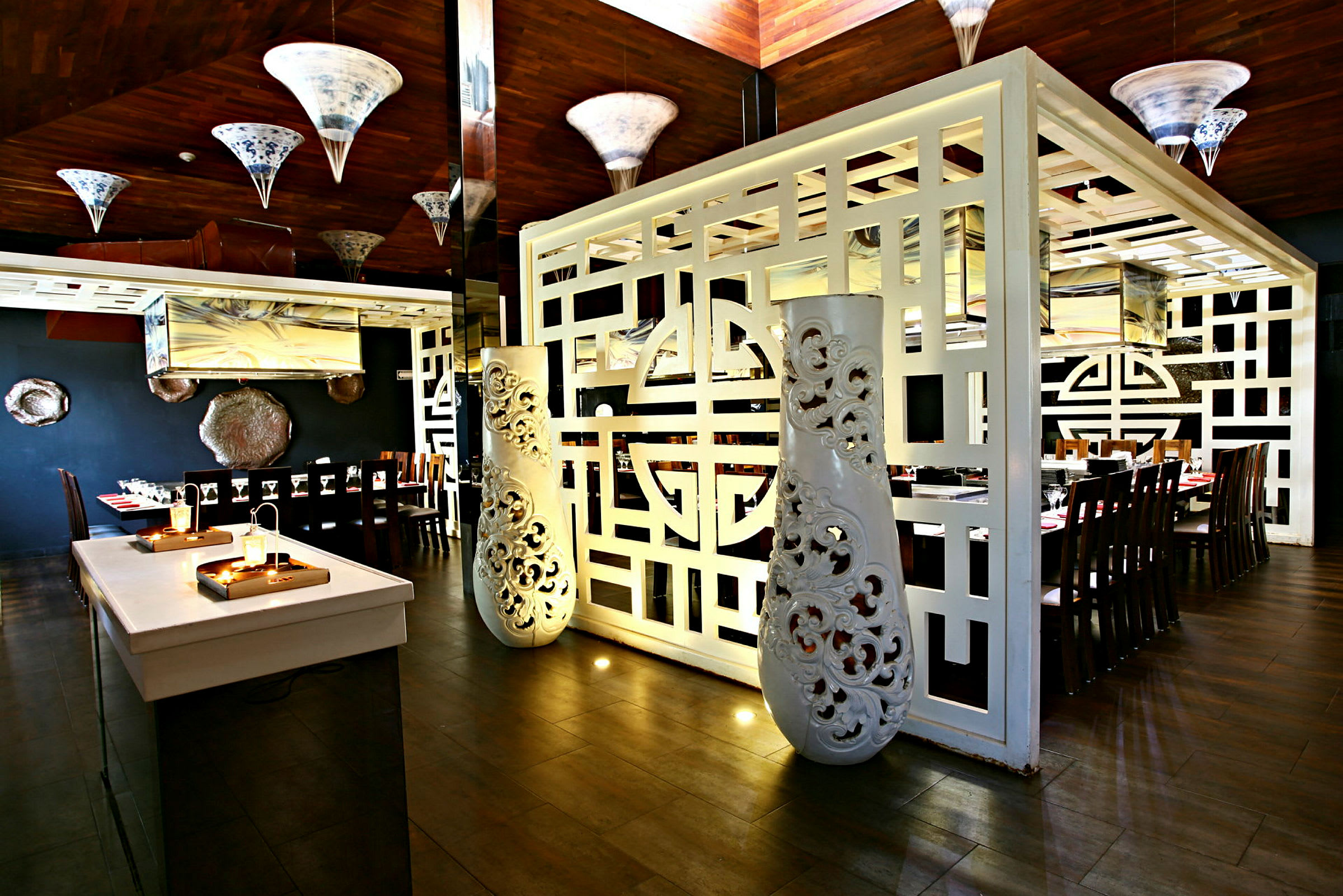En inomhusrestaurang med ett elegant inrett område med träbord och stolar. Rummet är dekorerat med vita ornamenterade skiljeväggar och hängande lampor. (AI-genererad text)