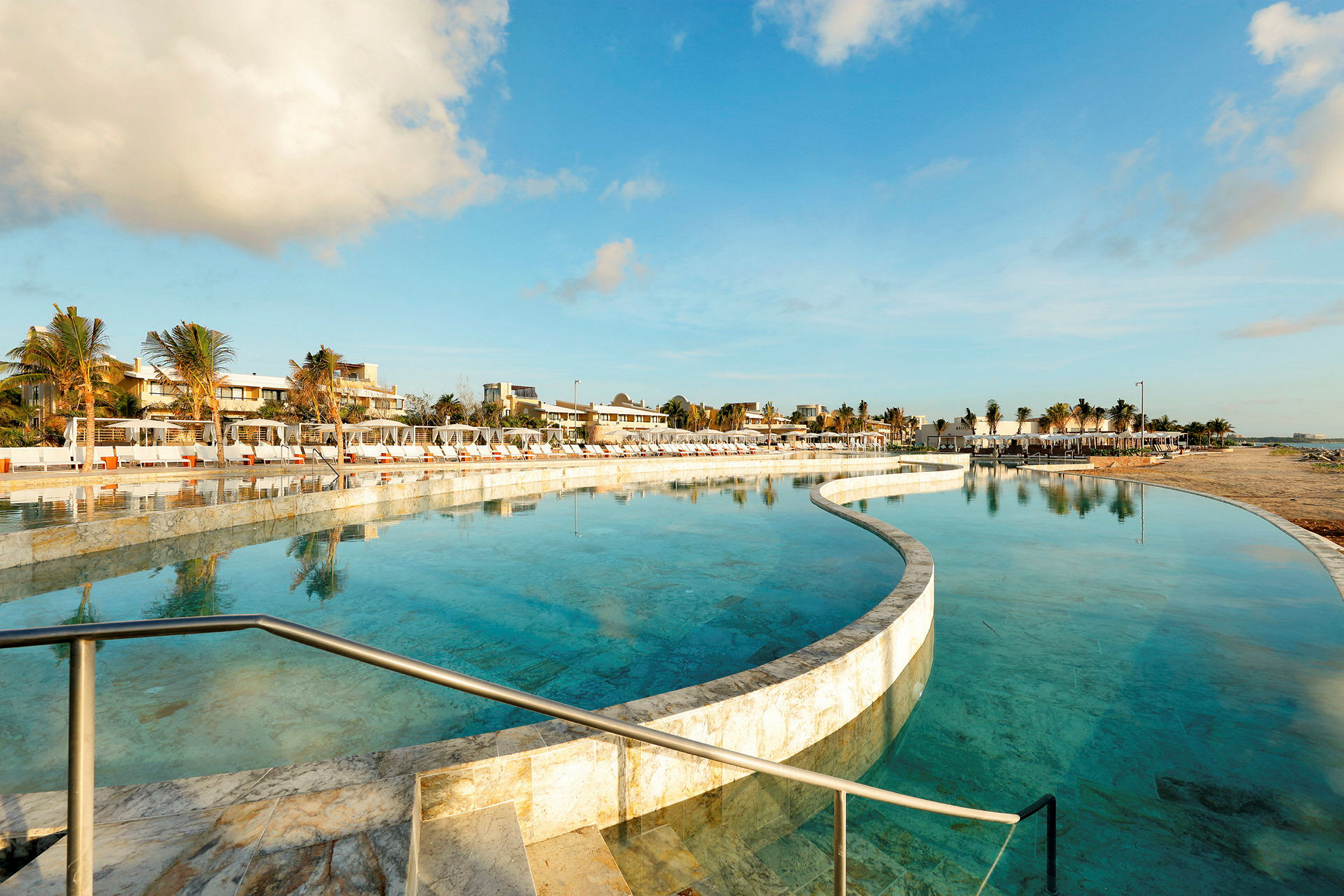 En strandnära pool omgiven av solstolar och palmer med klar himmel ovanför. (AI-genererad text)