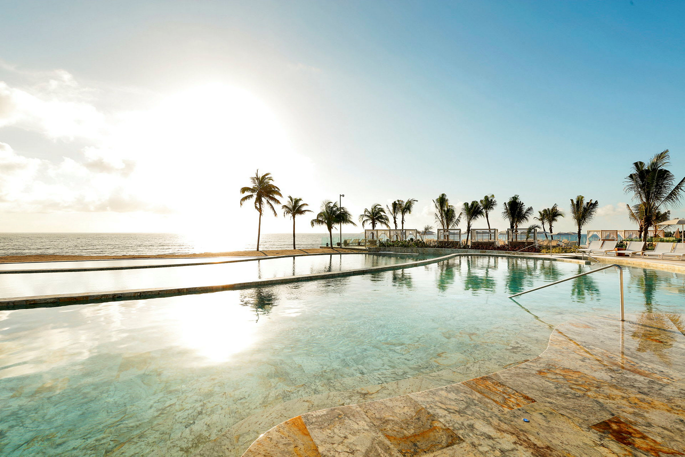 En infinitypool med klarblått vatten ligger framför en havsstrand kantad med palmer och solstolar. (AI-genererad text)