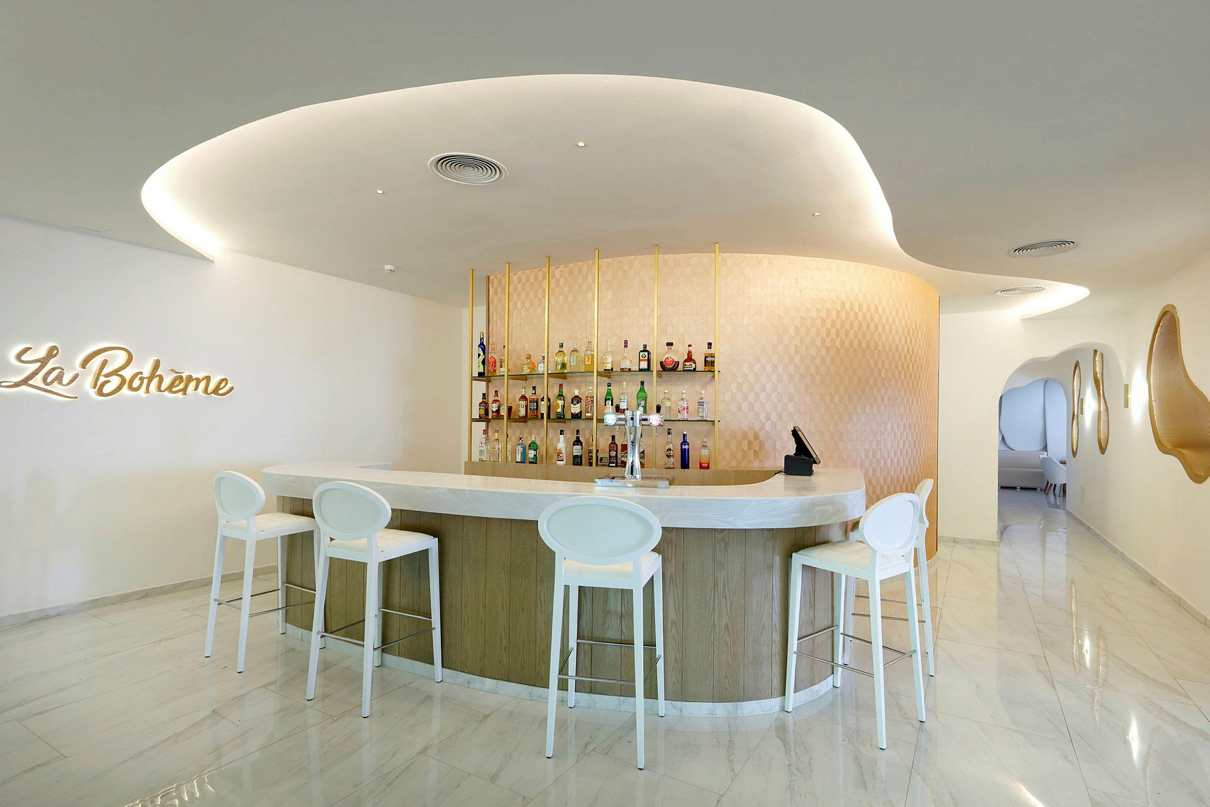 En modern bar med elegant inredning. Fyra vita barstolar står framför en böjd bardisk, och hyllor med flaskor syns bakom. Skylten 'La Bohème' är monterad på väggen. (AI-genererad text)