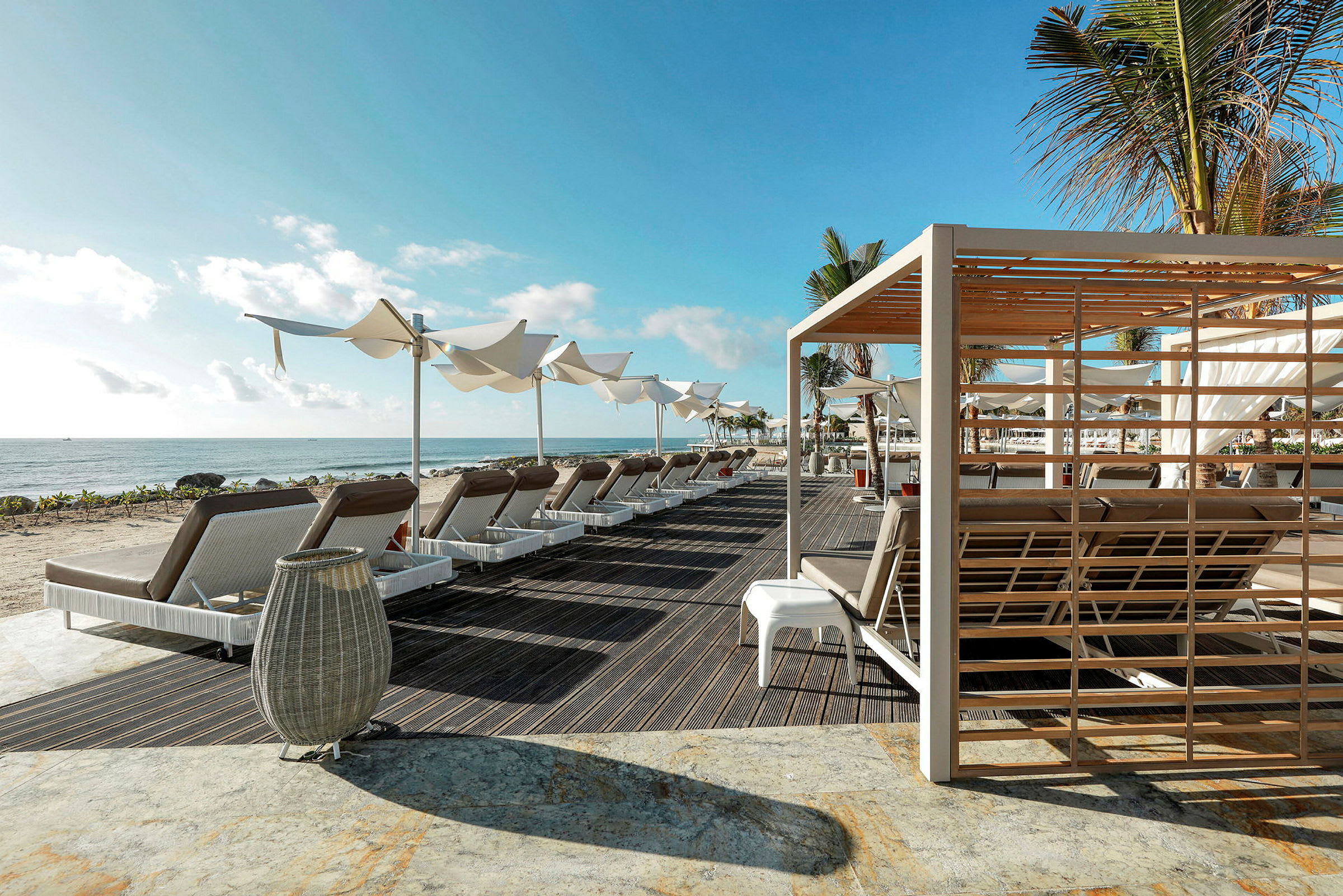 En strand med solstolar, parasoll och en pergola i förgrunden under en blå himmel. (AI-genererad text)