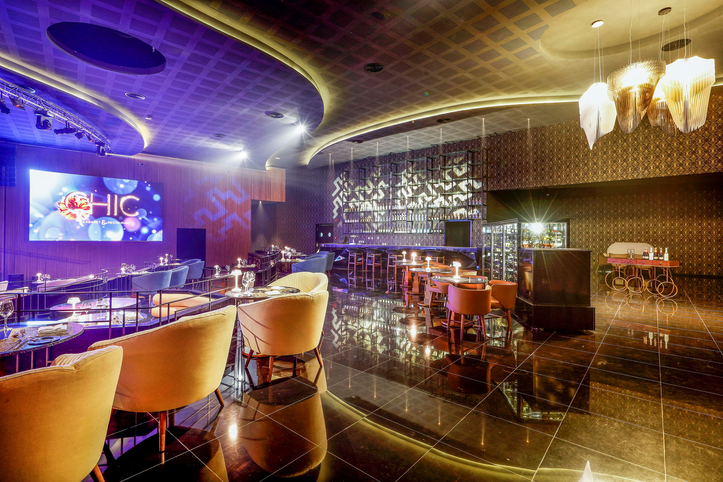 Ett elegant inrett bar- och restaurangområde med stämningsfull belysning, lyxiga sittplatser och en stor LED-skärm på väggen. (AI-genererad text)