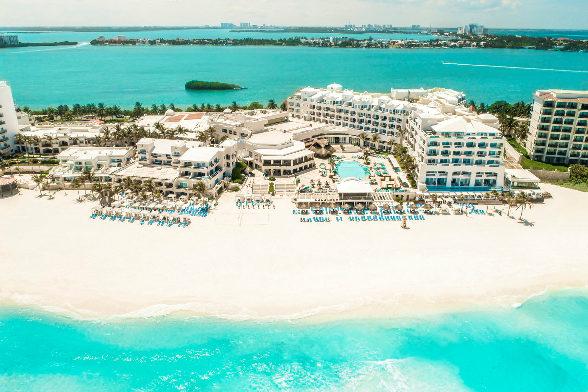 Flygfoto av hotellkomplex vid en vit sandstrand med turkost vatten och solstolar. (AI-genererad text)