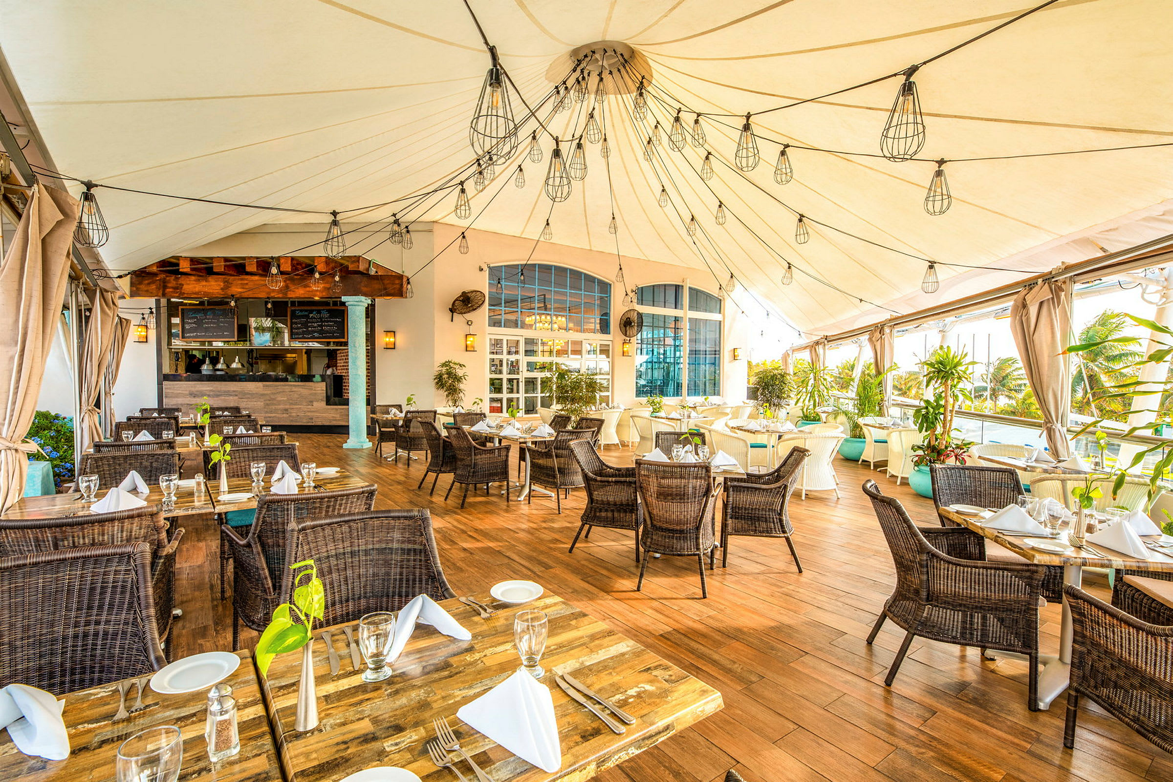 En restaurang med trägolv och dukade bord under ett tältliknande tak. Det finns gott om grönska och hängande lampor som dekoration. (AI-genererad text)