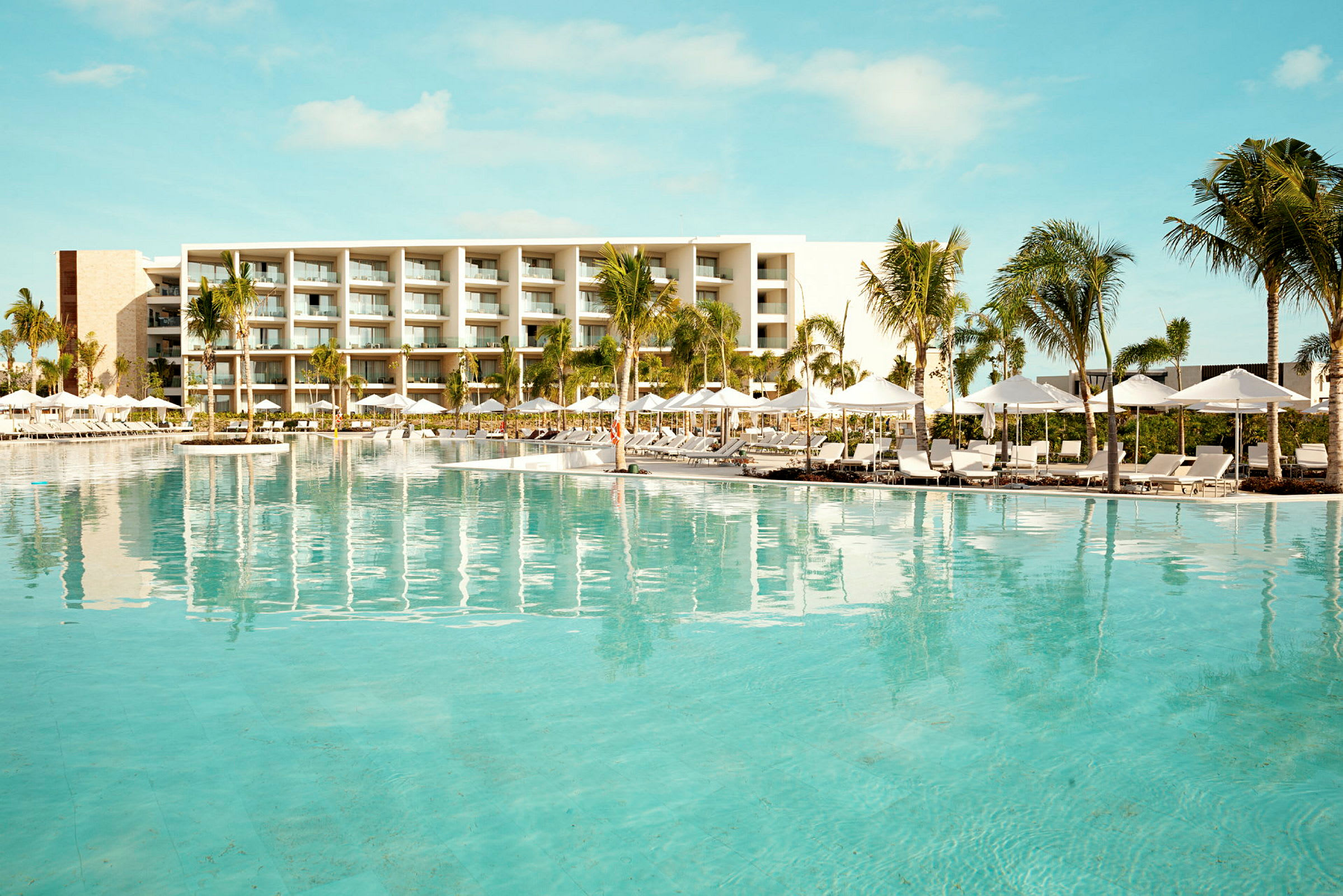 Bild av en hotellpool med solstolar och parasoller framför en modern byggnad. Palmträd omger området. (AI-genererad text)