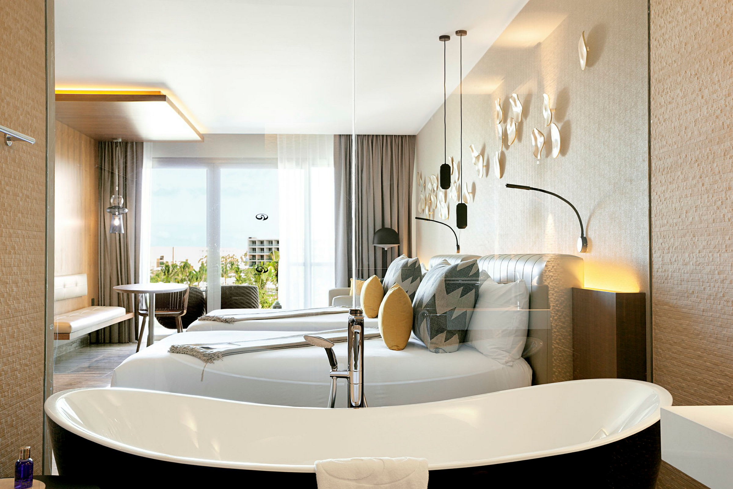 Ett modernt hotellrum med en stor dubbelsäng, dekorativa kuddar och väggdekorationer. Det finns ett fristående badkar och möbler såsom bord och stolar nära en glasdörr med utsikt. (AI-genererad text)