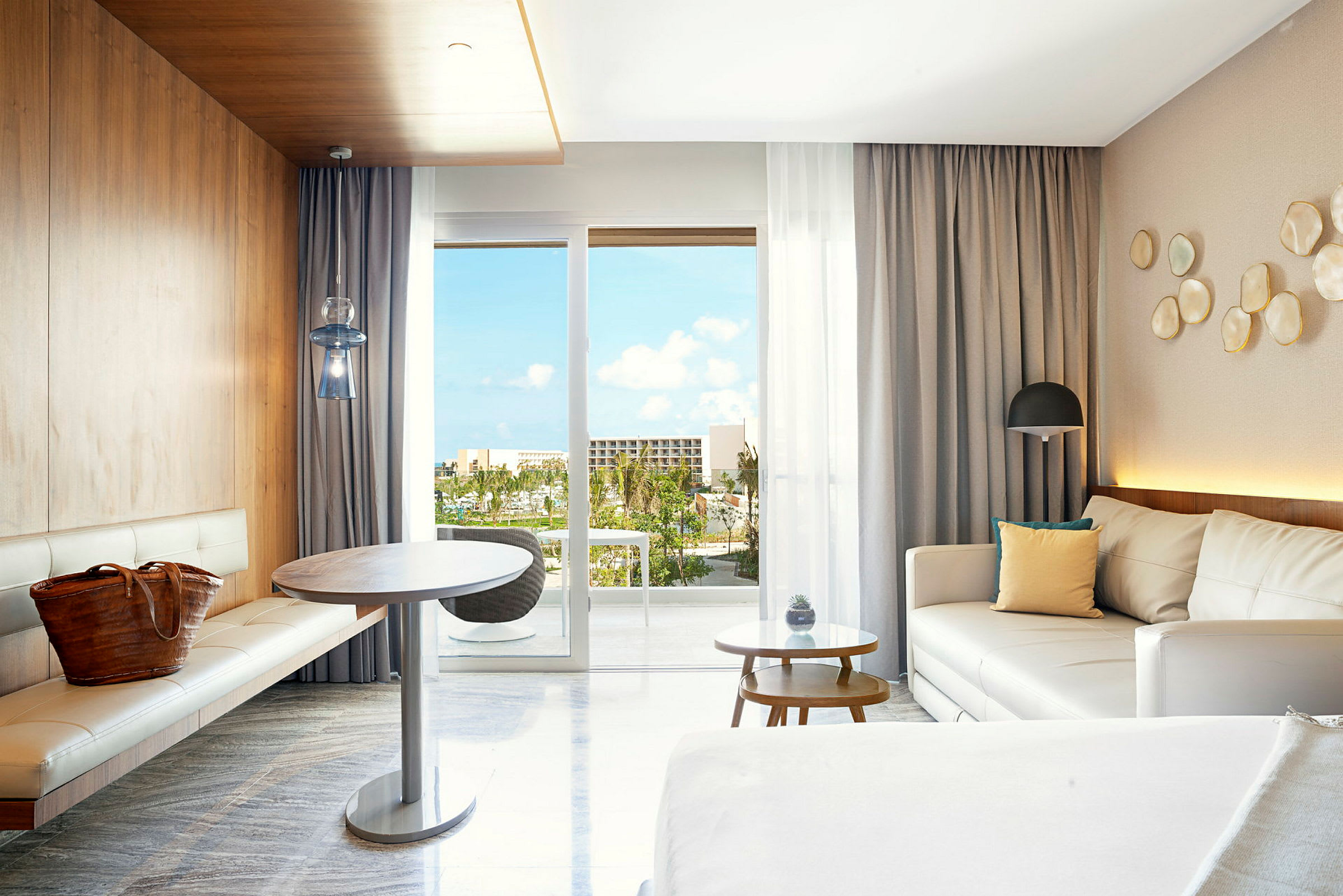 Ett modernt hotellrum med vita möbler och träpaneler. Utsikt över balkongens panoramafönster med tropiska växter och blå himmel synlig. (AI-genererad text)
