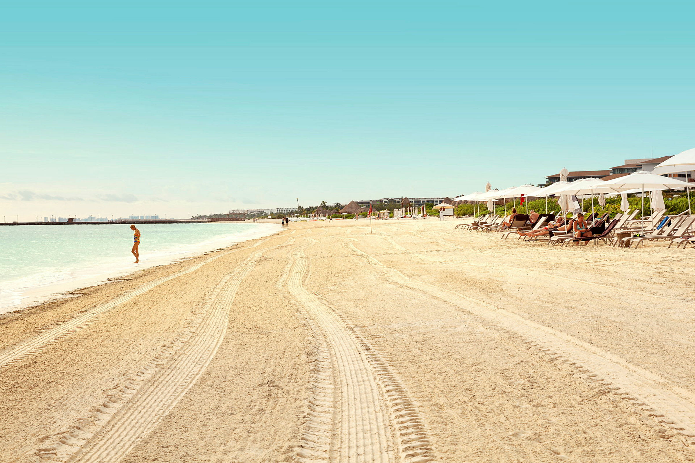 En sandstrand med parasoller och solstolar, en person står vid vattnet. (AI-genererad text)