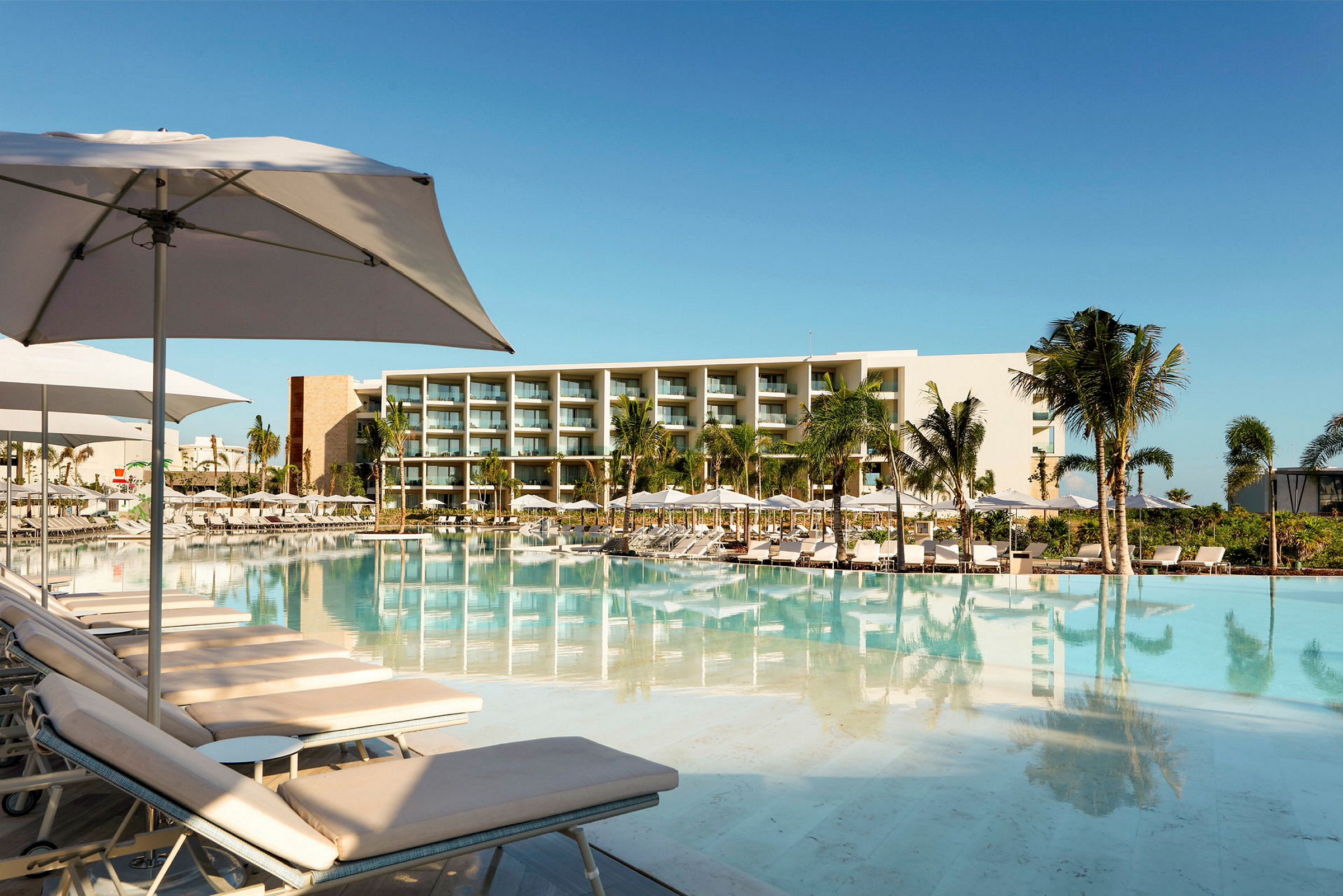 Hotell i modern stil med stor pool, omgiven av solstolar och parasoller samt palmträd. (AI-genererad text)