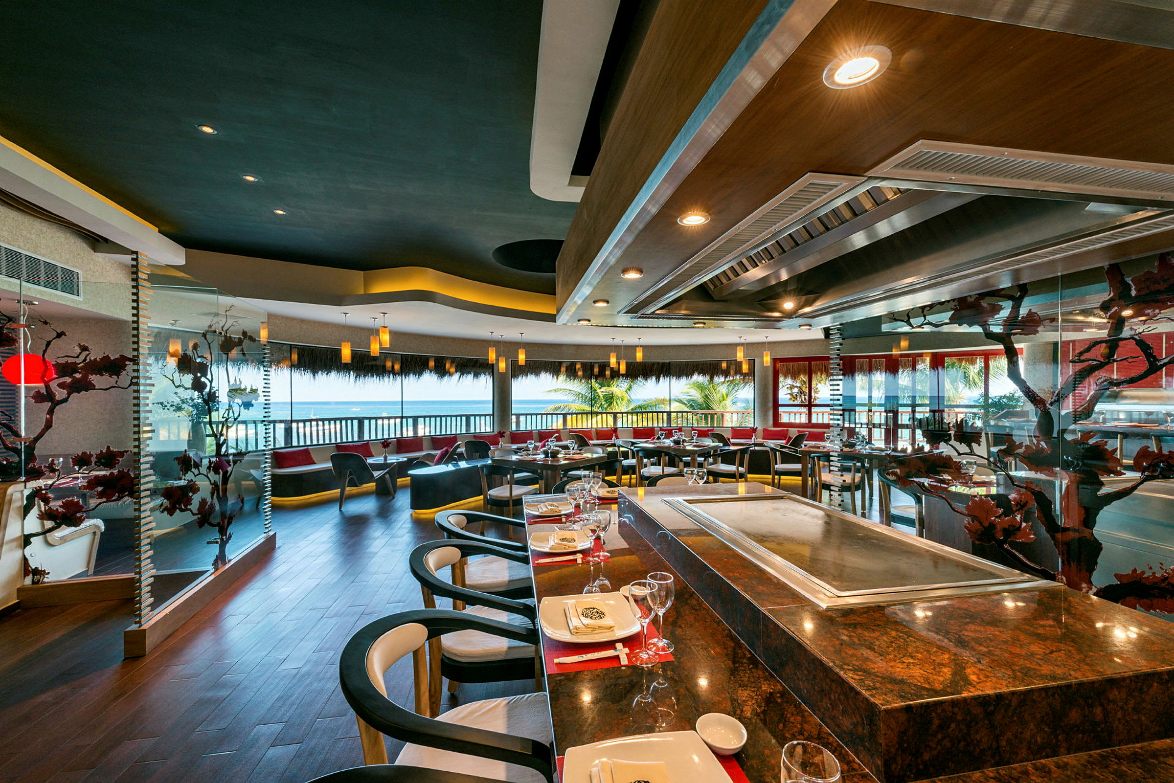 En modern restaurang med bord och stolar, stora fönster med utsikt över palmer och en grillstation i förgrunden. (AI-genererad text)