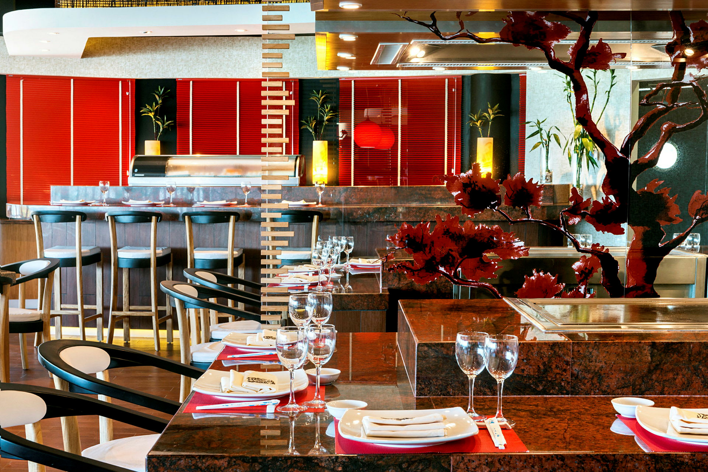 Ett stylat restauranginteriör med bord dukade med glas och tallrikar samt stolar. En bar med stolar syns i bakgrunden, och lokalen har dekorativa detaljer och ljus inslag. (AI-genererad text)