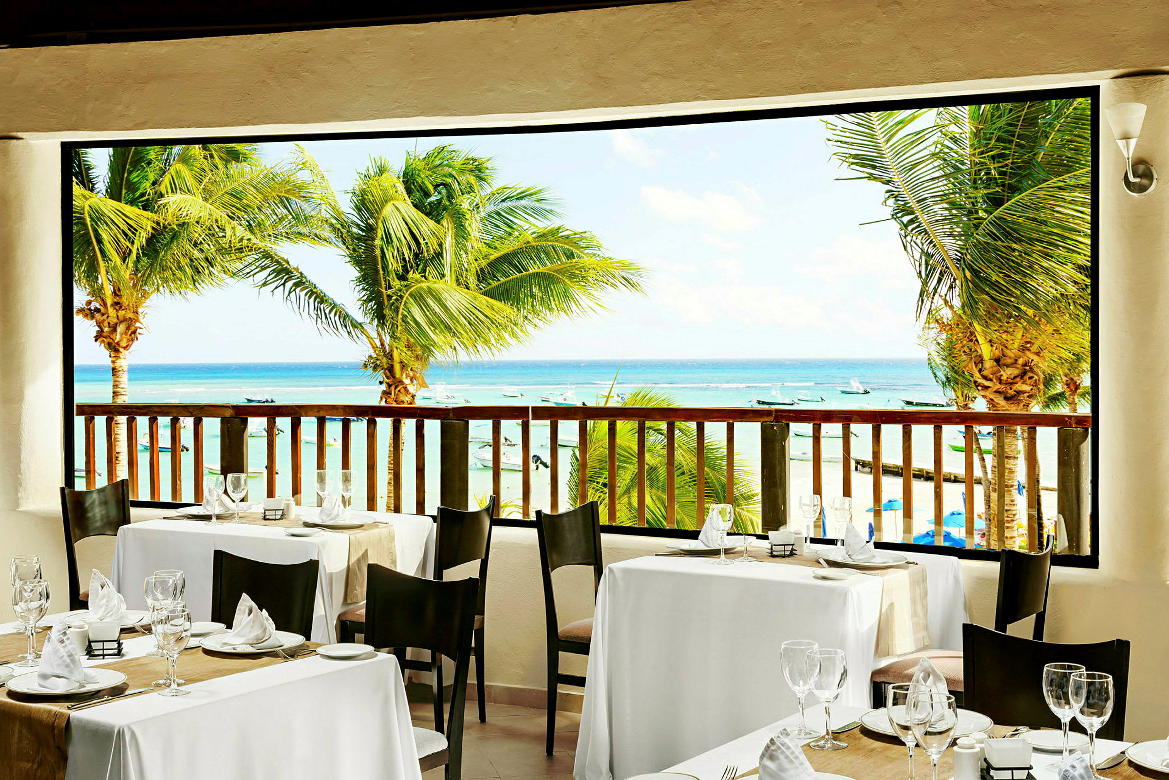 Utsikt från en restaurang med dukade bord mot stranden och havet, omgiven av palmer. (AI-genererad text)
