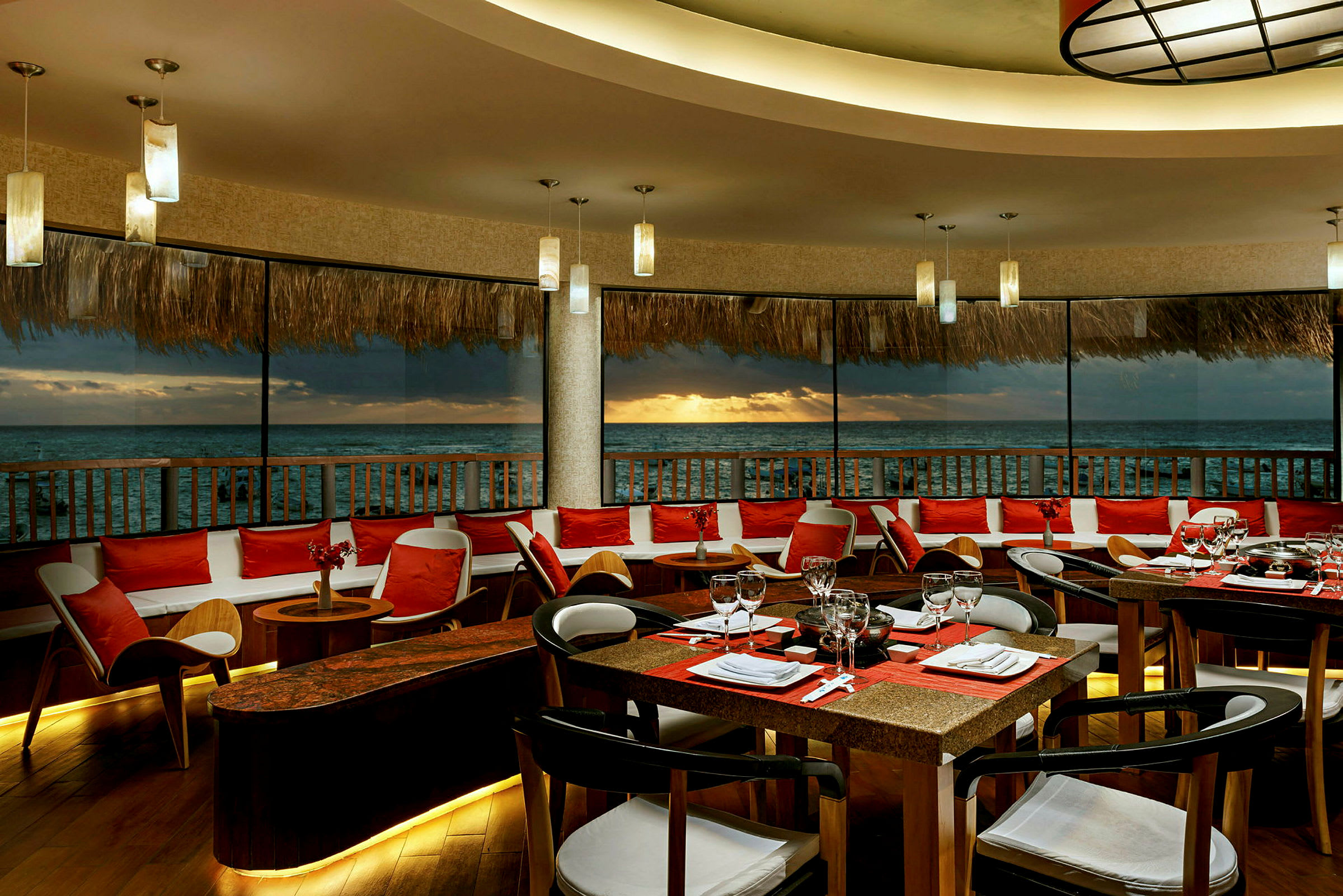 En restaurang med bord och stolar placerad vid ett stort fönster med utsikt över havet och solnedgången. (AI-genererad text)