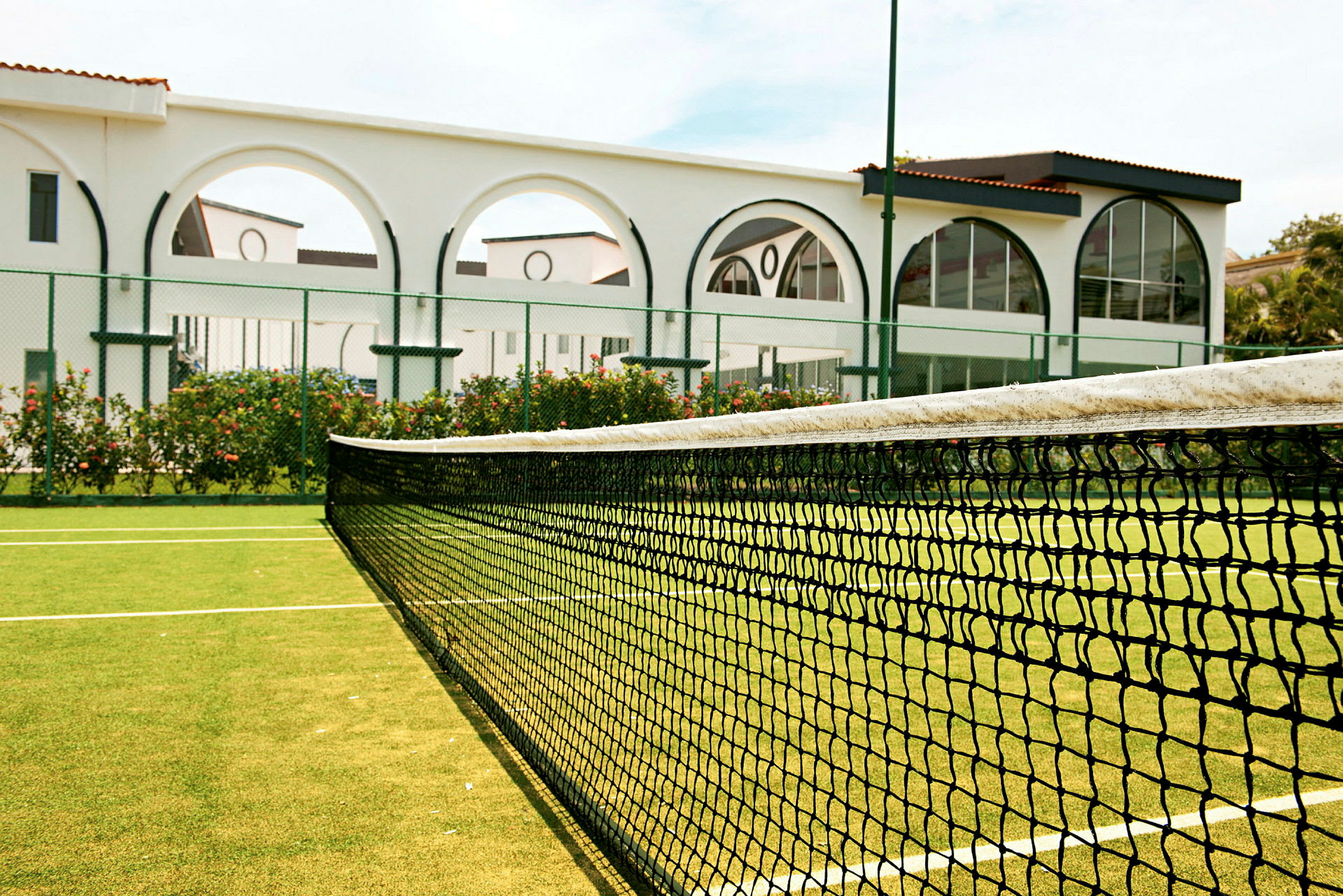 En tennisplan med ett nät i förgrunden. Bakom nätet syns en vit byggnad med valv och glaspartier. (AI-genererad text)