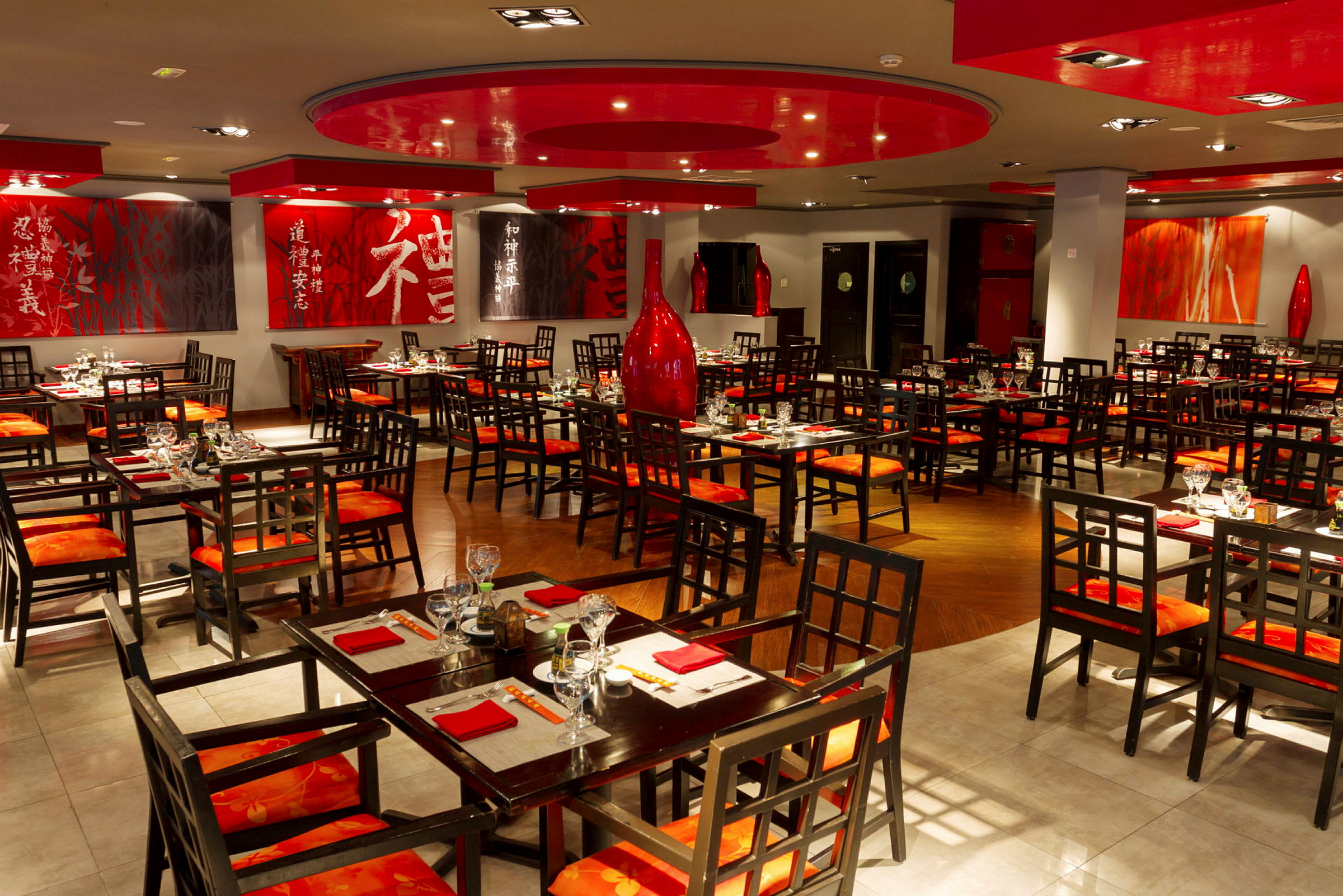 En restaurang med asiatisk inredning och uppsättningar av bord och stolar i trä. Väggarna är dekorerade med konst och röda accenter. (AI-genererad text)