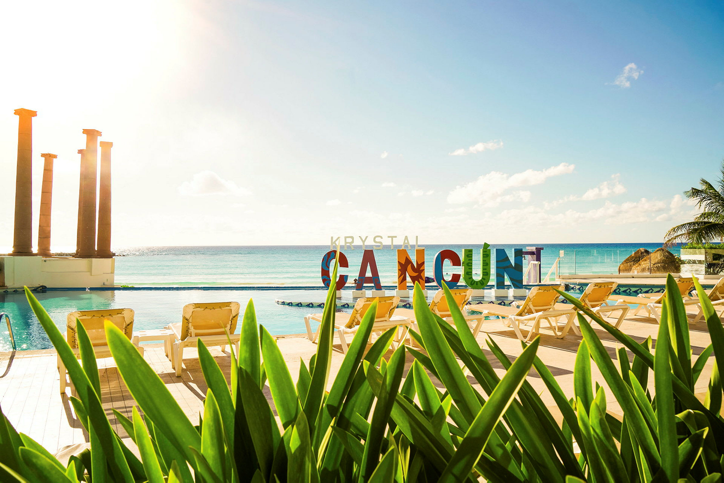 Utsikt över en pool med solstolar och grönska i förgrunden. I bakgrunden syns havet och färgglada bokstäver som stavar "CANCUN". (AI-genererad text)