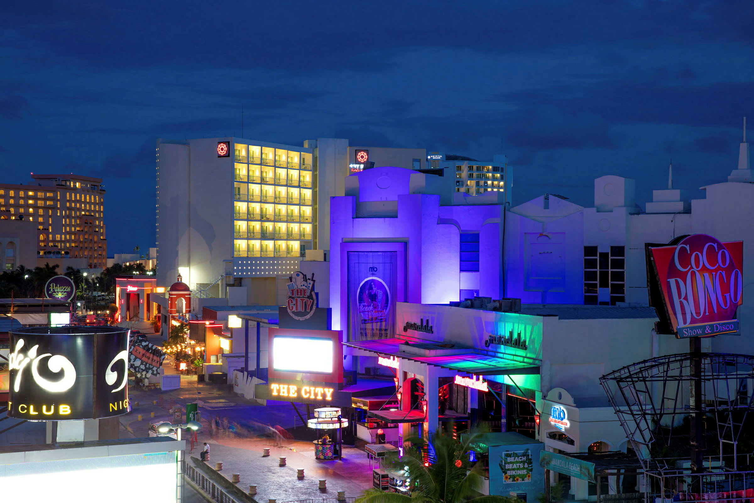 Bild av ett nattklubbsområde med färgglada byggnader och skyltar, inklusive en tydlig skylt för Coco Bongo. Bakgrunden visar hotell med upplysta fönster. (AI-genererad text)