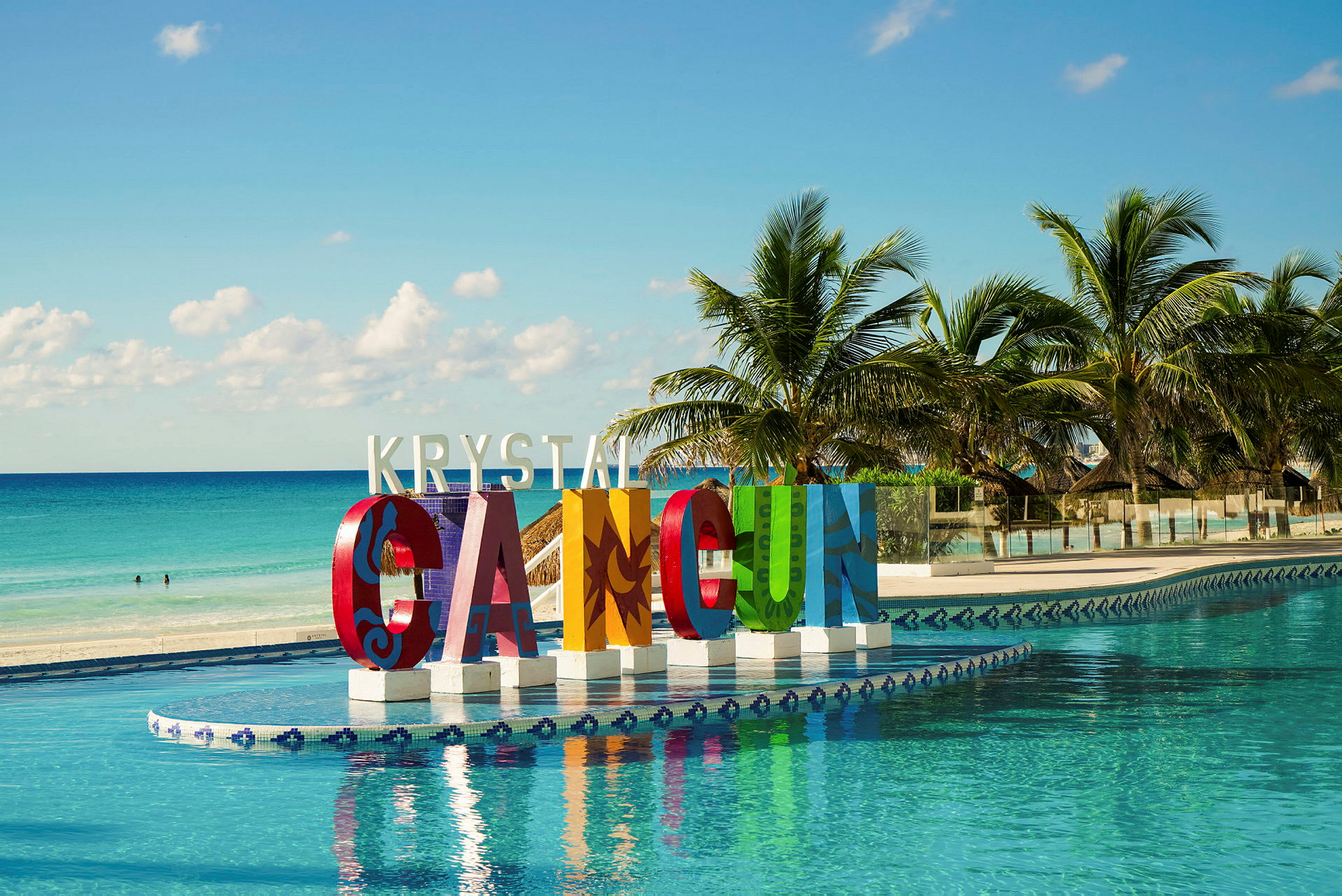 Stora färgglada bokstäver som bildar texten 'Cancun' vid en pool. Palmer och hav i bakgrunden. (AI-genererad text)