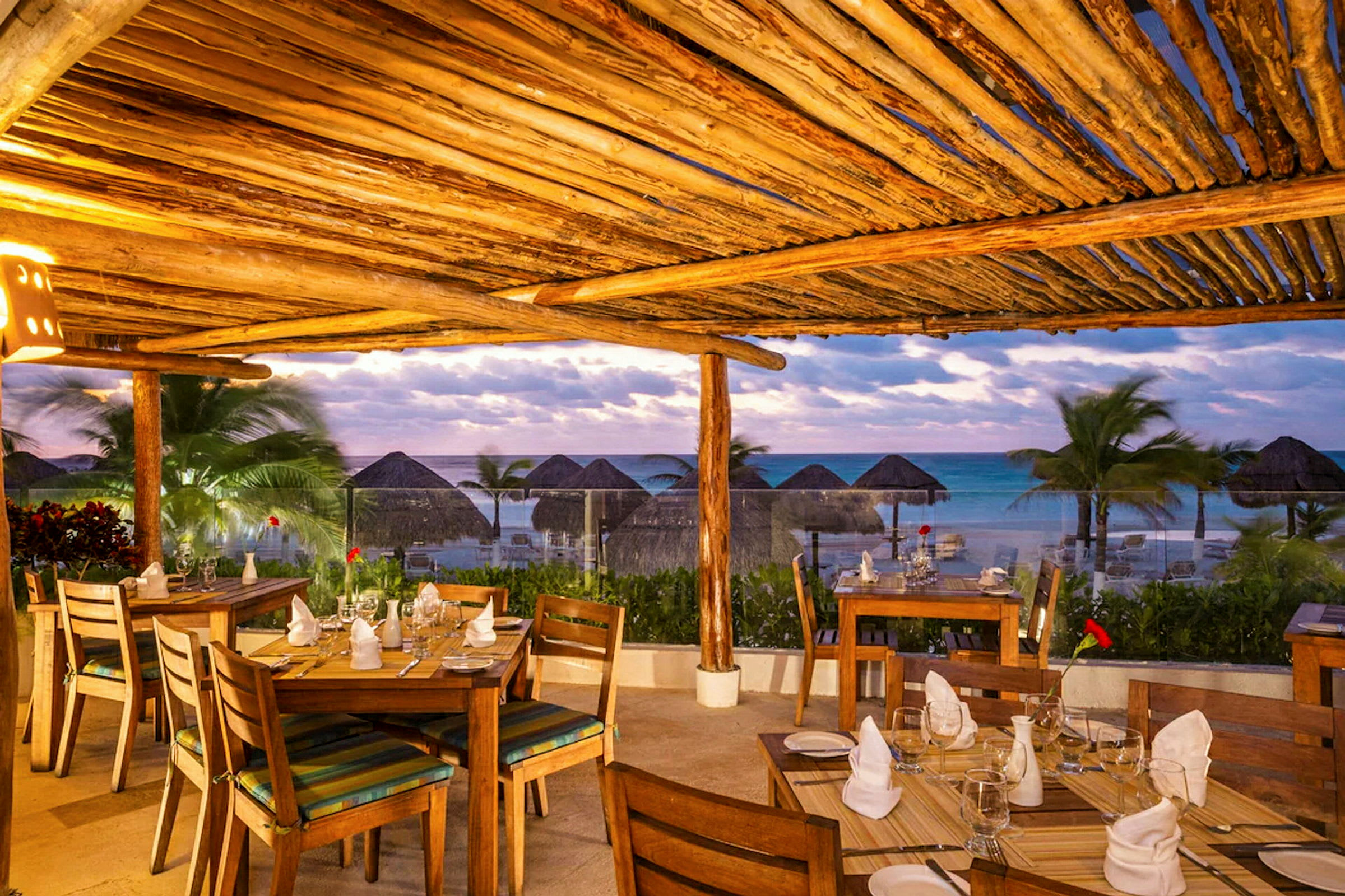 Restaurang med träbord och stolar under ett tak av trä med utsikt över havet och palmer. (AI-genererad text)