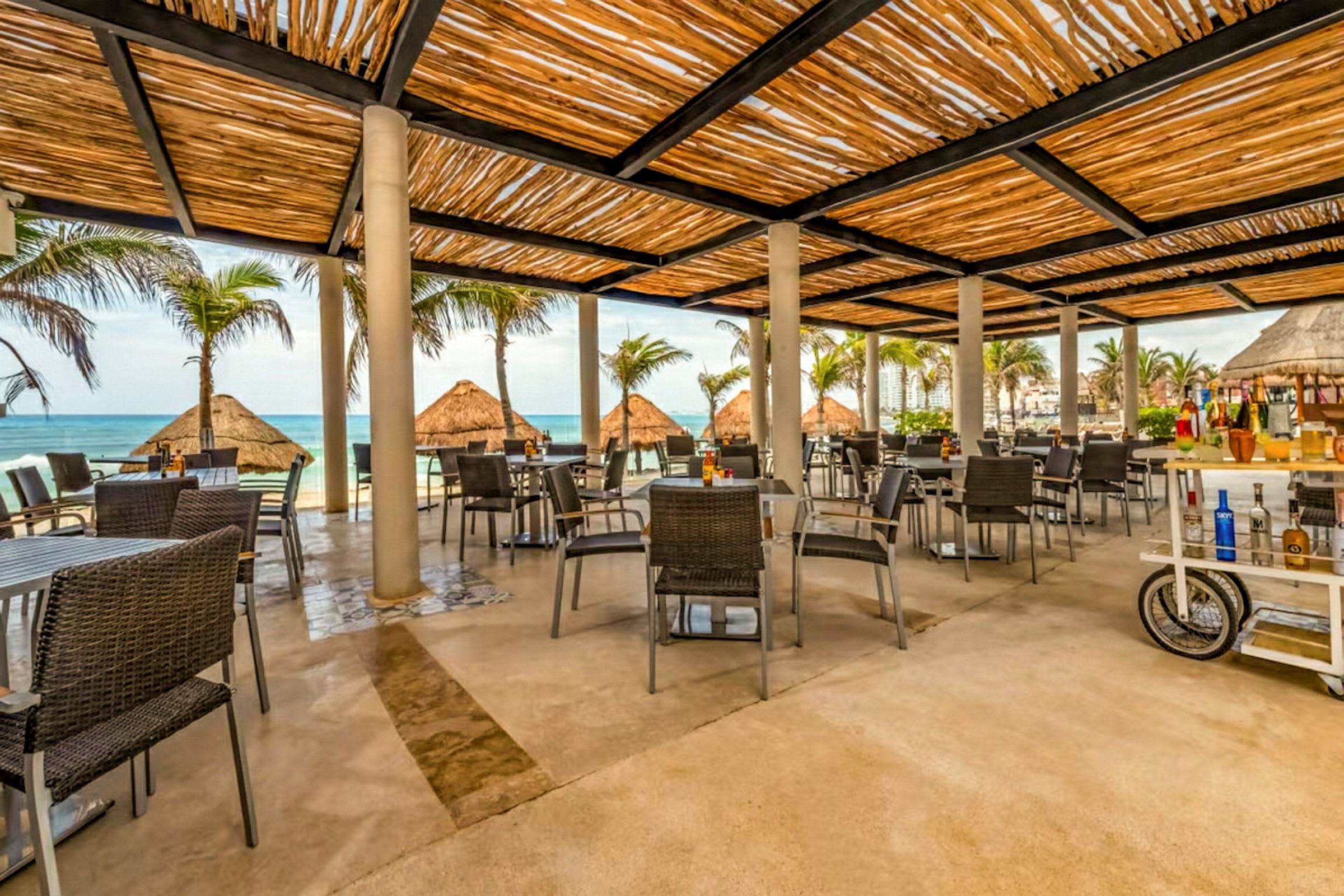 En utomhusrestaurang med bord och stolar under ett tak av träribbor, med utsikt över stranden och havet. (AI-genererad text)