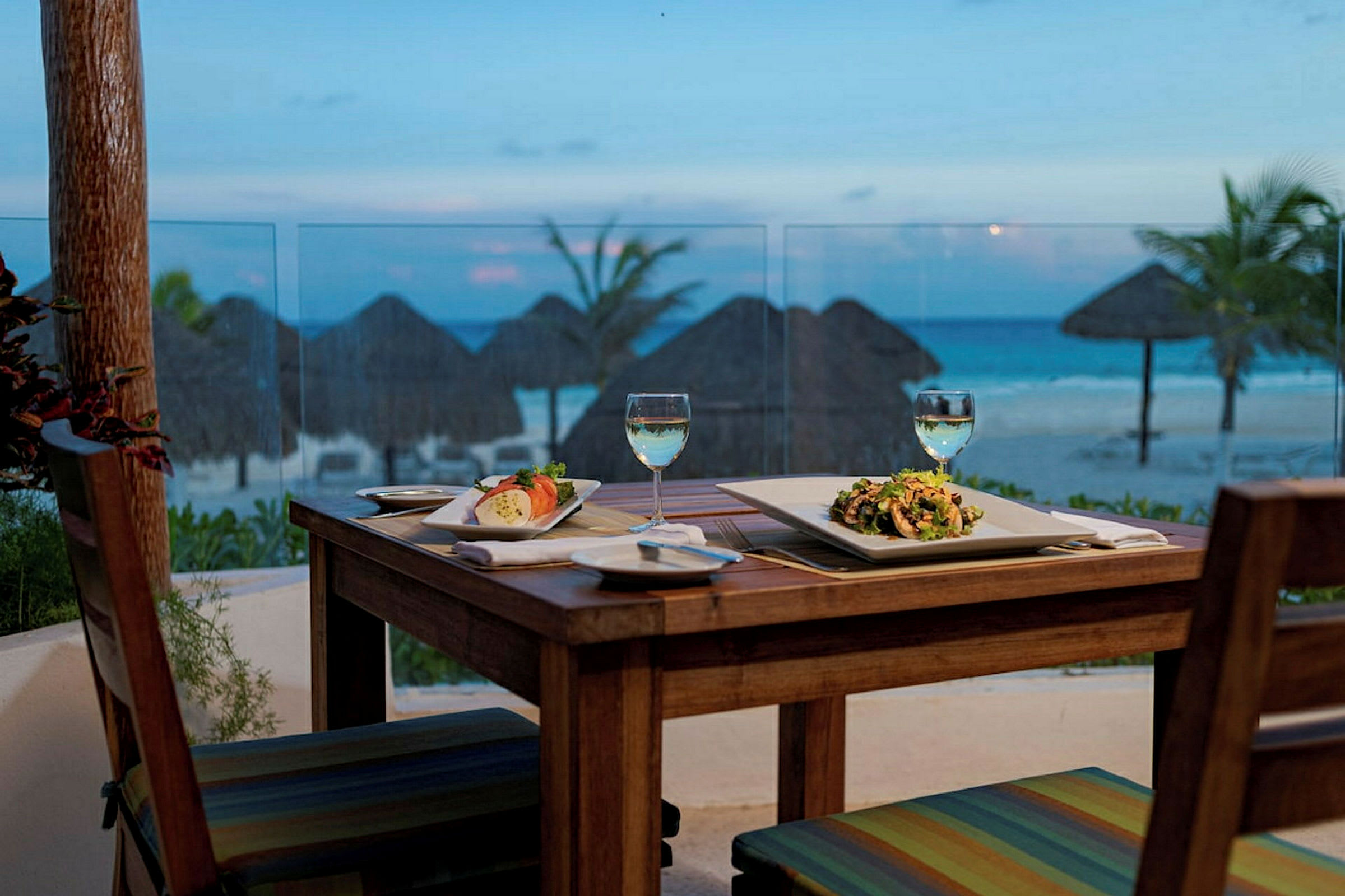 Ett dukat bord vid en uteservering med två tallrikar med måltider och två glas vin, med utsikt över en strand och havet. (AI-genererad text)
