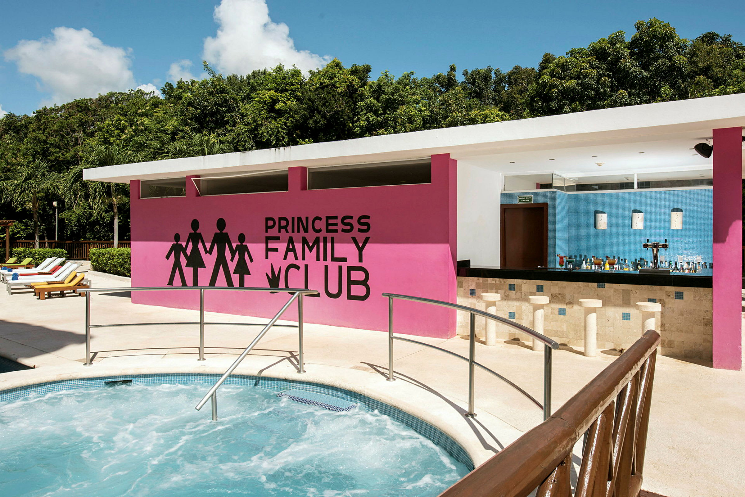 En utomhuspool med en jacuzzi i förgrunden, en rosa vägg med texten 'Princess Family Club' och en bar under ett tak. Det är omgiven av grönska och solsängar finns vid sidan. (AI-genererad text)