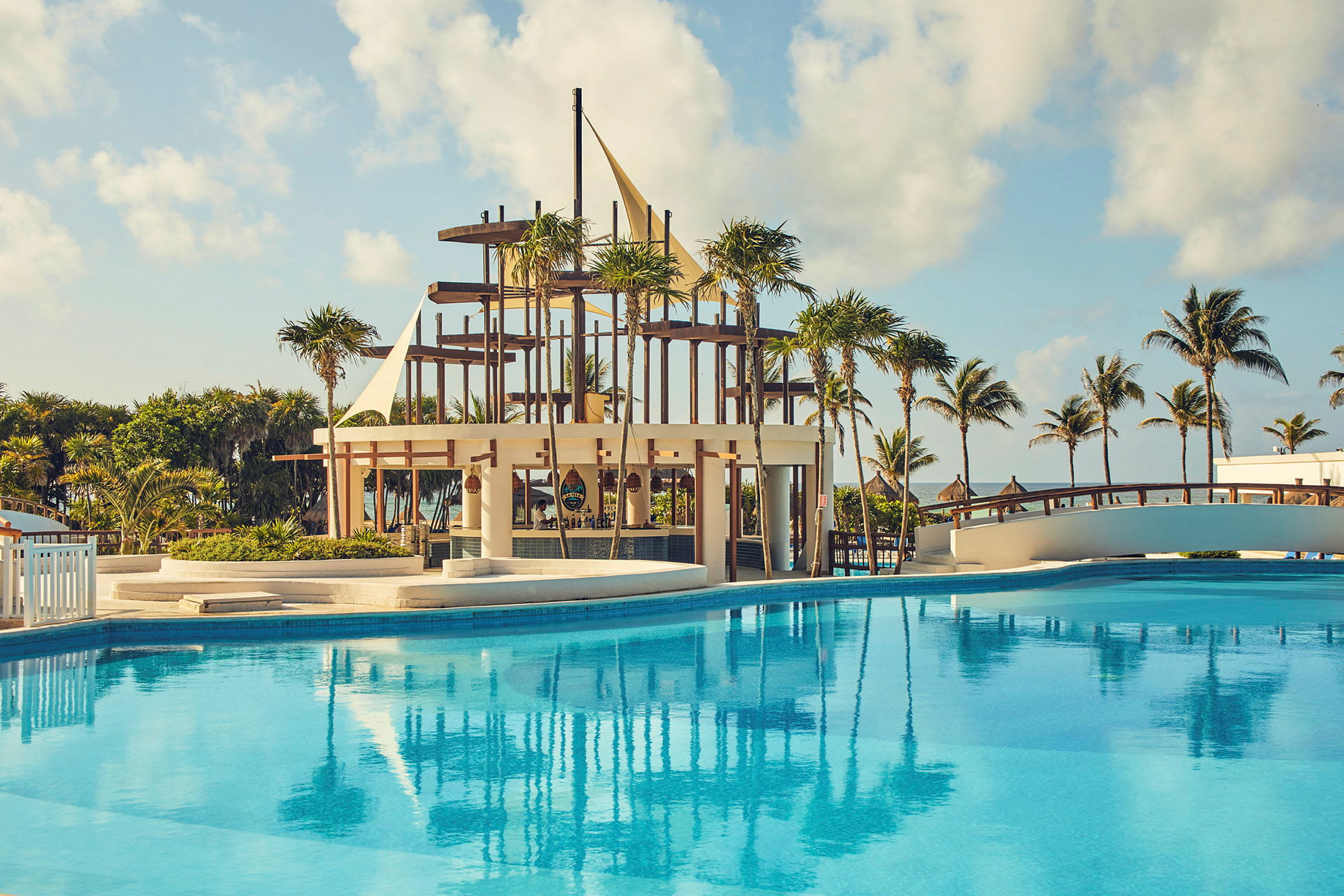 En pool med klarblått vatten framför en strandbar med konstruktioner av trä och segelliknande detaljer. Palmträd och en bro syns i bakgrunden, samt en klar himmel. (AI-genererad text)