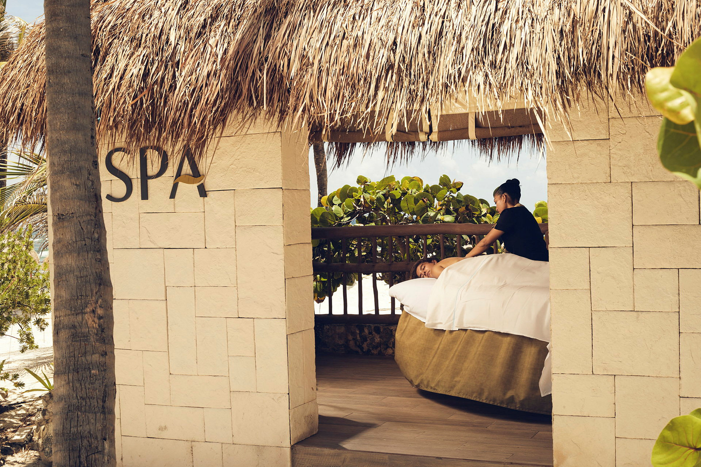 En utomhus spa-paviljong under ett halmtak, där en person får en avslappnande massage. (AI-genererad text)