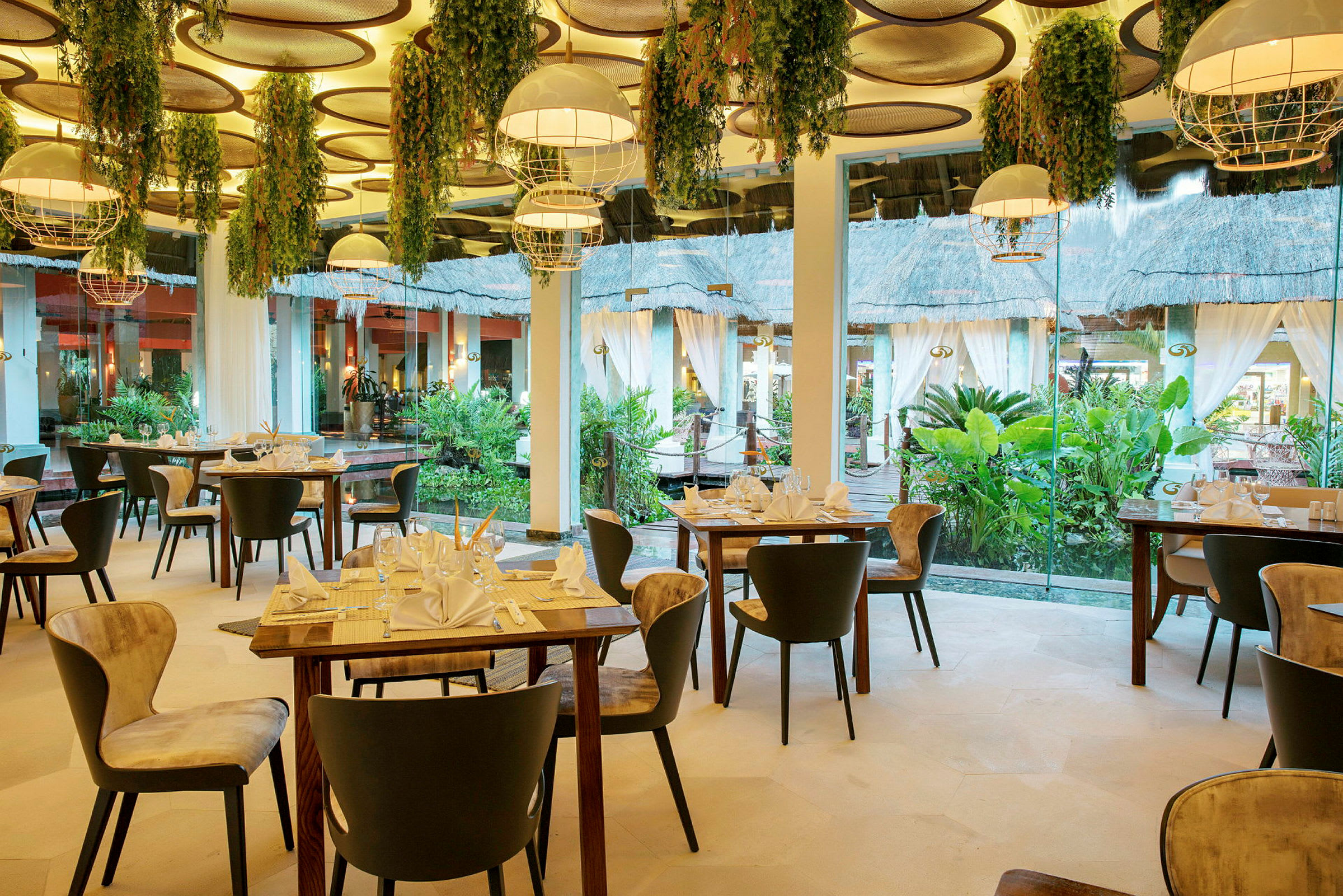Interiör av en elegant restaurang med dekorerade bord och stolar, hängande lampor och växter, samt stora fönster som vetter mot en lummig trädgård. (AI-genererad text)
