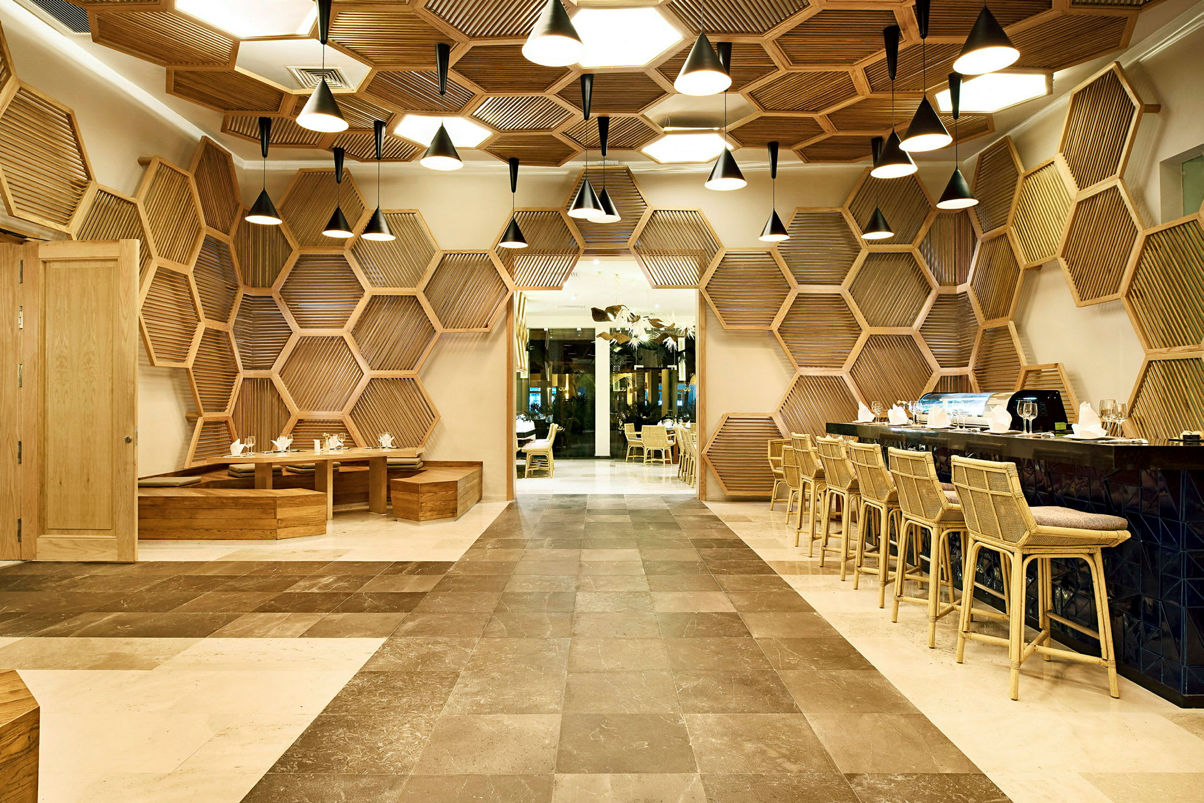 En restaurang med modern design, hexagonformade träpaneler på väggarna och taket och eleganta ljuskronor. Bord och stolar i trä finns på två sidor. (AI-genererad text)