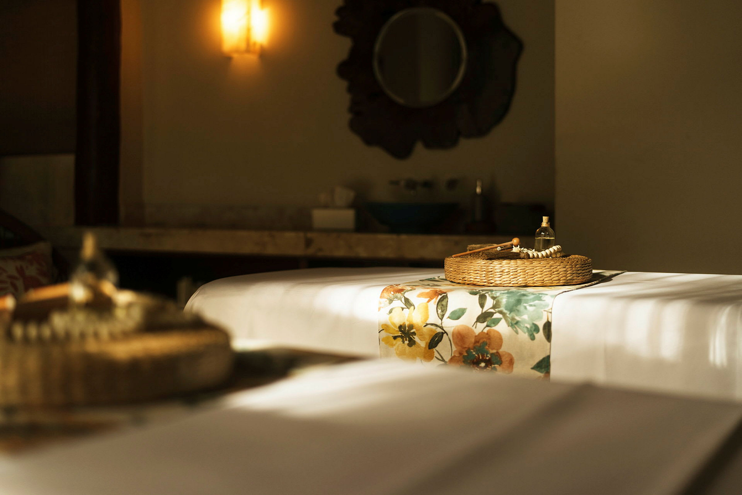 Ett rum med massagebord klätt i vitt tyg och blommigt överdrag, med en korg innehållande en glasflaska och massageverktyg. En spegel och belysning syns i bakgrunden. (AI-genererad text)
