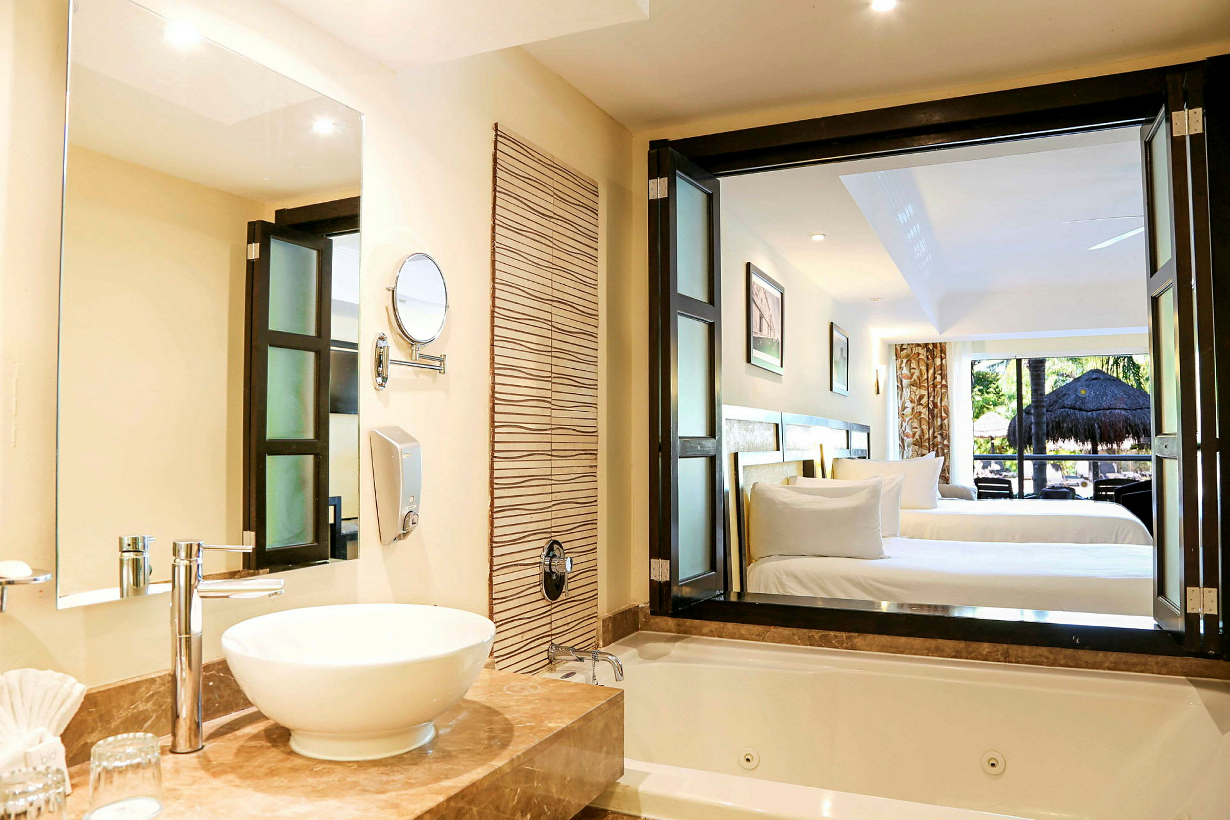 Ett hotellrum med badrum i förgrunden som har ett tvättställ på ett marmorbord och en jacuzzi. I bakgrunden syns en sovdel med två enkelsängar och en vy över en balkong med grönska. (AI-genererad text)