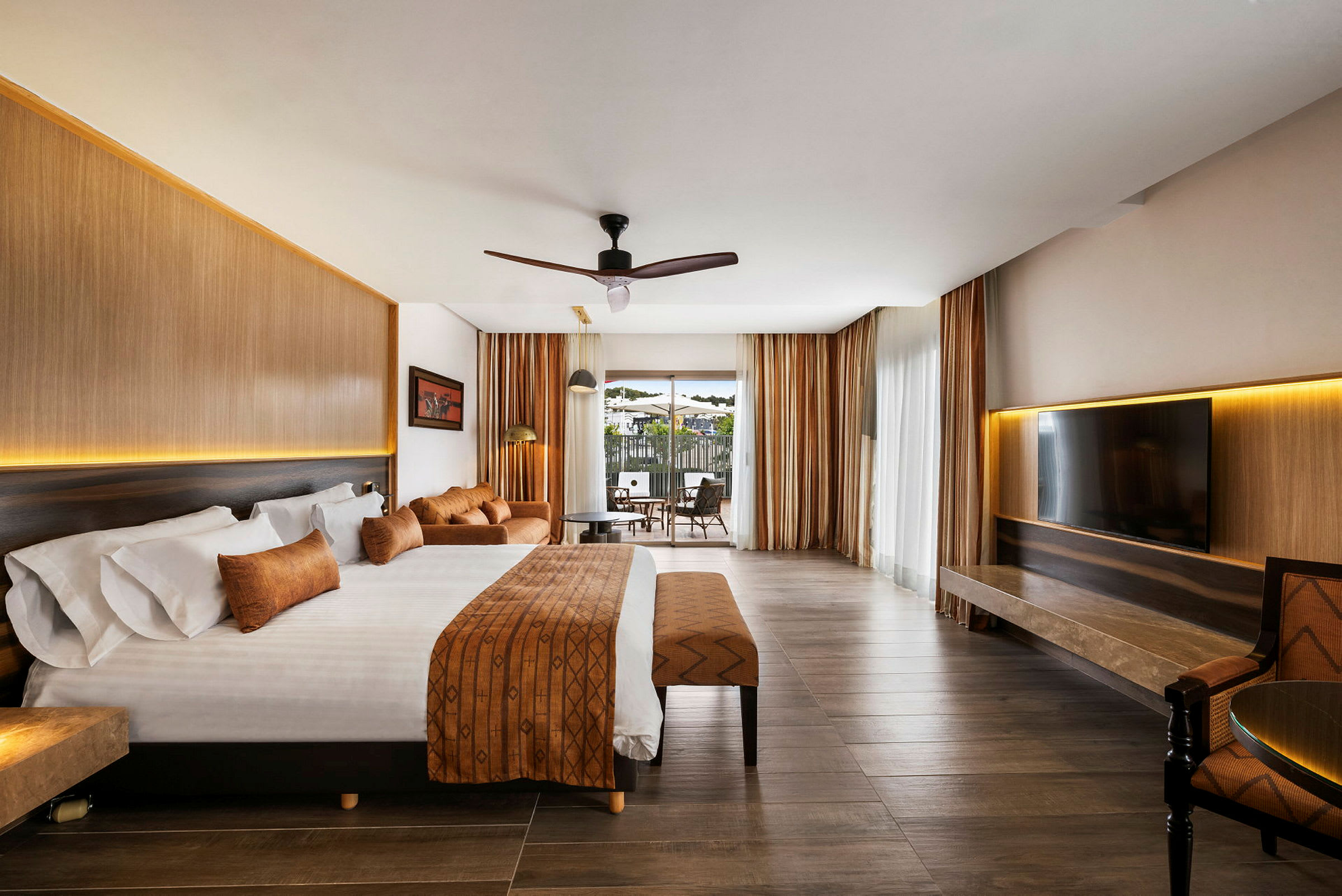 Hotellrum med stor säng, sittgrupp och utsikt genom glasdörrar. (AI-genererad text)