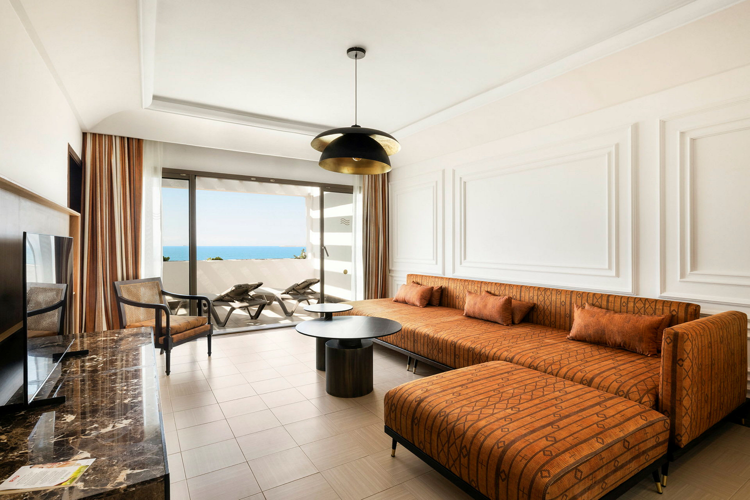 Ett modernt hotellrum med en orange soffa, ett soffbord och utsikt mot havet från en balkong. (AI-genererad text)