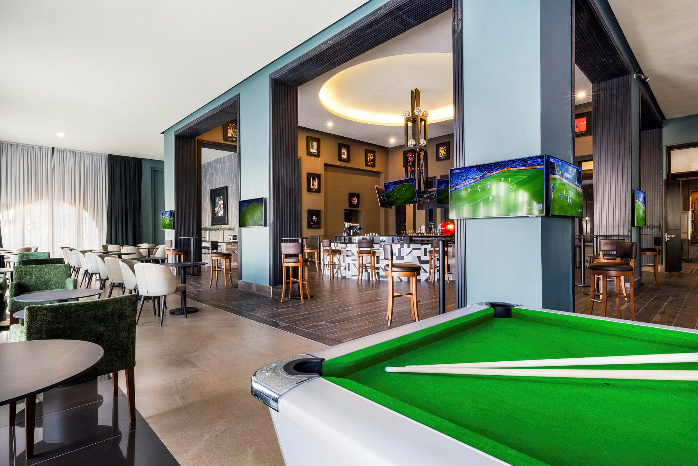 Interiör av en modern lounge med sittgrupper, ett biljardbord och flera TV-skärmar som visar sport. (AI-genererad text)